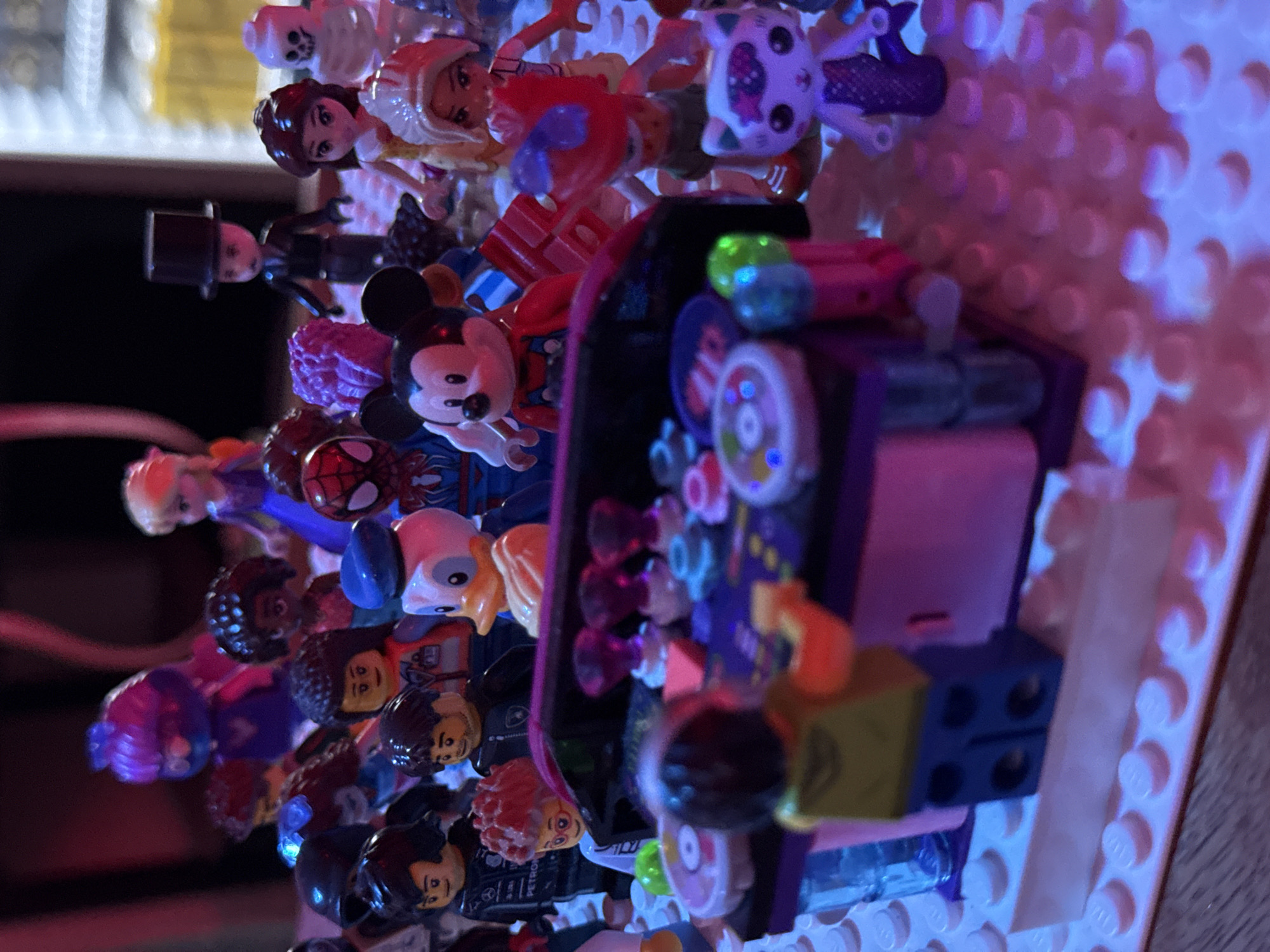 The image shows a collection of LEGO minifigures arranged on a light pink LEGO baseplate, illuminated by purple and blue lights. In the foreground, a minifigure with a yellow shirt and blue pants stands behind a DJ booth with purple and pink accents. The DJ booth has several colorful knobs and lights.
Surrounding the DJ booth are other LEGO minifigures, including Mickey Mouse, Donald Duck, Spider-Man, Elsa, a skeleton figure, Belle, and a figure with a top hat. There are also several other minifigures whose identities are less clear due to the lighting and focus. The background is dark, with hints of a blurred yellow and white structure. The scene appears to be indoors, with the LEGO creations set up on a wooden surface. The location in Si Sunthon, Thailand, is not discernible from the image.