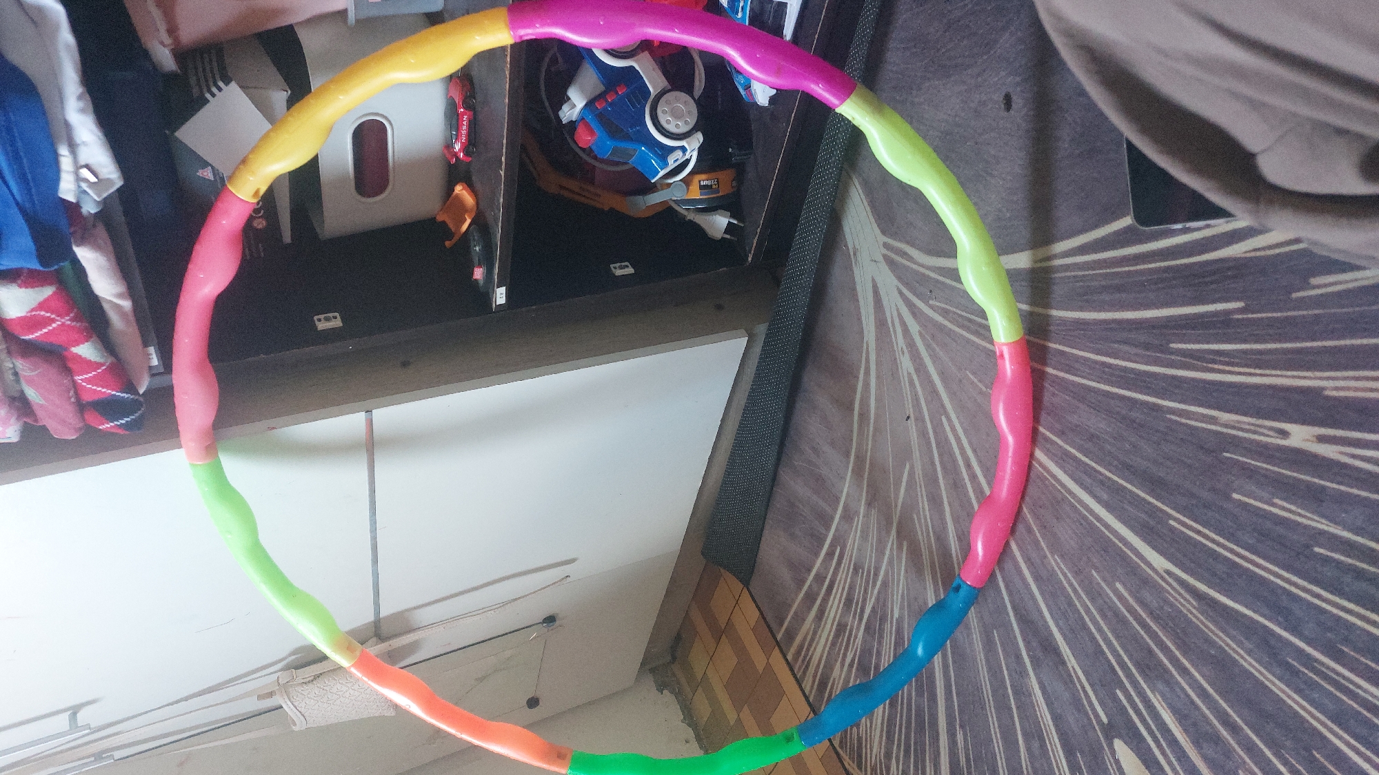 A multi-colored hula hoop, constructed of interlocking sections in pink, blue, yellow, green, and orange, lies partially on a patterned rug and partially on a dark mat. The hula hoop is the primary subject, positioned in the foreground and midground of the image. Behind the hula hoop is a dark-colored shelving unit filled with various items. Visible on the shelves are folded clothes, a white storage bin, a toy red Nissan car, a toy blue truck, and other indistinct objects. To the left of the shelving unit, a portion of a light-colored wall is visible, with a light-colored woven bag hanging from a thin strap. The floor is covered by a brown and beige patterned rug. The scene appears to be an indoor domestic setting, possibly a room in a home. There is no indication of a specific location within the city of Jatirogo, Indonesia. No individuals are visible in the image.