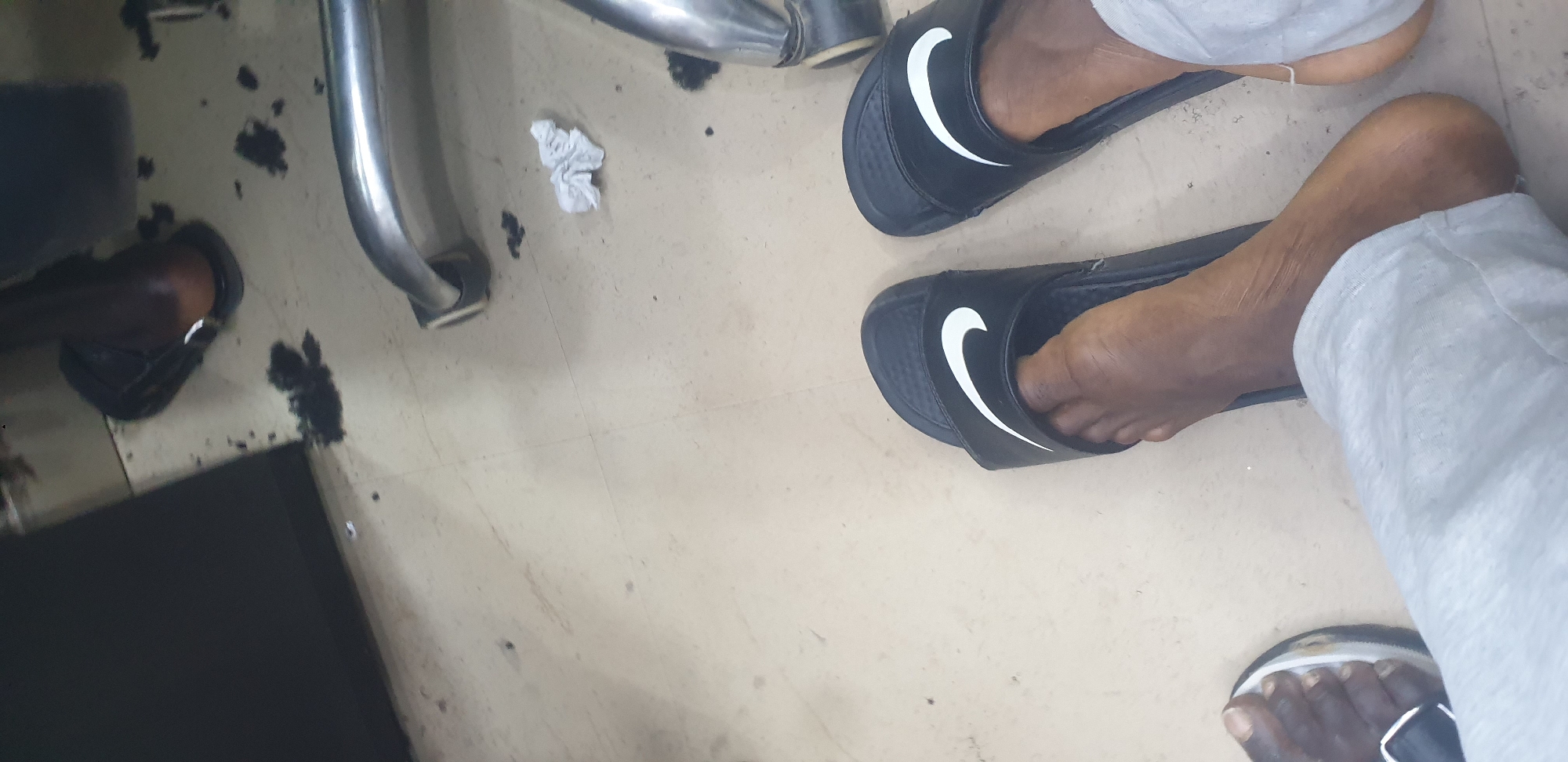 The image depicts a ground-level view of what appears to be a barbershop floor. Two individuals' feet are visible, both wearing black Nike slides with white swooshes. One foot on the right shows a portion of light-colored pants. The floor is tiled and appears to have scattered dark clippings, consistent with a haircutting environment. A crumpled white tissue is also visible on the floor. In the background, a metallic chair leg and a dark, possibly upholstered, surface are present. The presence of hair clippings suggests this scene is likely in Jos, Nigeria, a common location for such businesses.