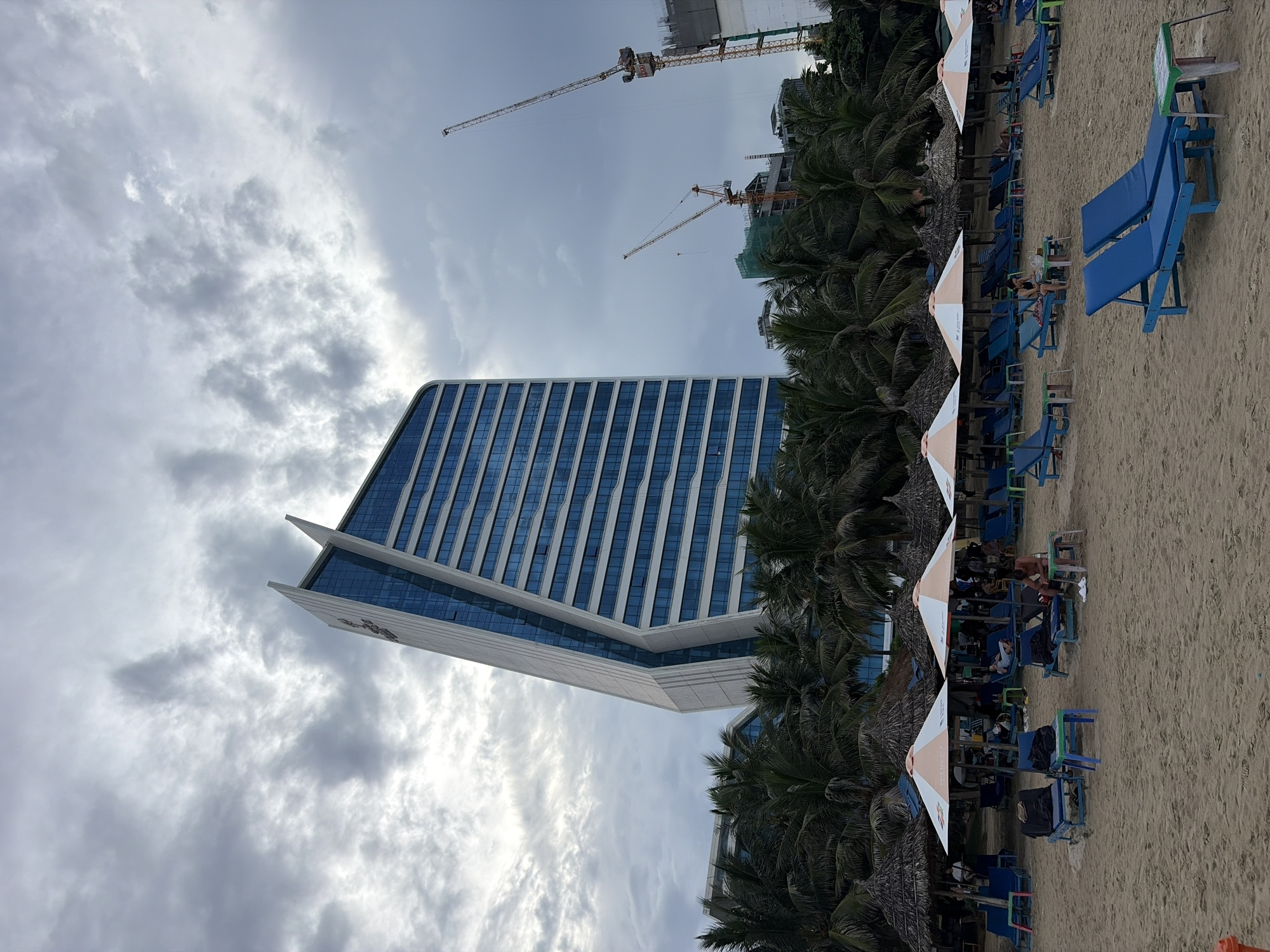 The image depicts a beach scene in Đà Nẵng, Vietnam, dominated by the distinctive architecture of the Ariyana Smart Condotel Đà Nẵng. In the background, the strikingly angled, modern building with its blue-tinted windows stands against a cloudy sky. Palm trees line the edge of the beach, forming a dense green backdrop to the scene.
The foreground is a sandy beach dotted with numerous lounge chairs and thatched umbrellas, suggesting a resort or public beach area. Several blue lounge chairs are unoccupied, while others are occupied by people relaxing. Some individuals are seated, and one person is shown at a small table. There are also several small, colorful tables placed near the lounge chairs.
Further in the background, beyond the palm trees and hotel, construction cranes are visible, indicating ongoing development in the city. The overall atmosphere is one of a cloudy day at a beachside resort, with both leisure activities and urban development present.
