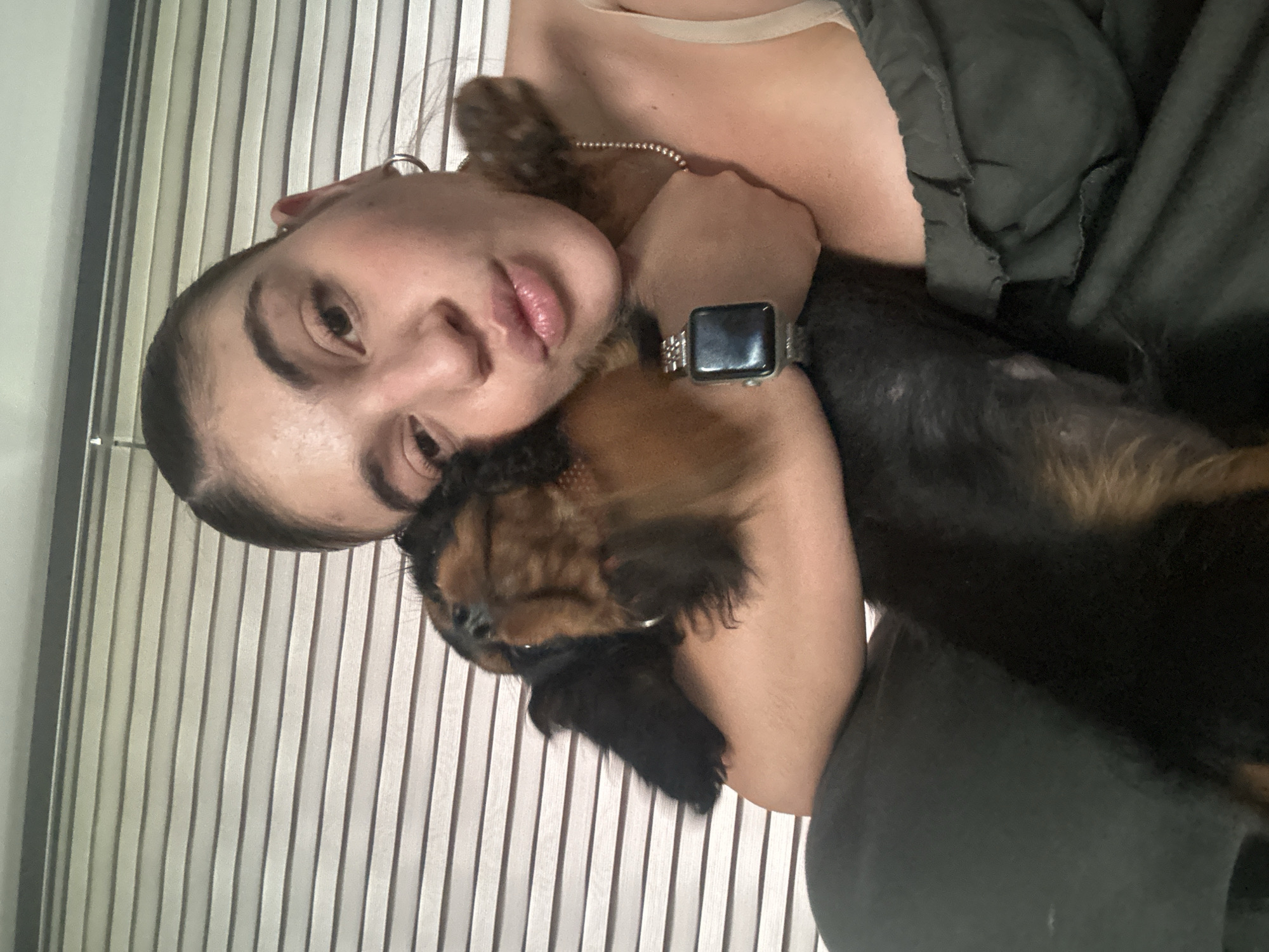 A woman with fair skin and dark eyebrows is hugging a small dog with black and brown fur. The woman is wearing an Apple Watch and a silver necklace. The background appears to be a set of vertical blinds. The location is unknown, but the question specifies Puebla, Mexico, though no details in the image support this location.