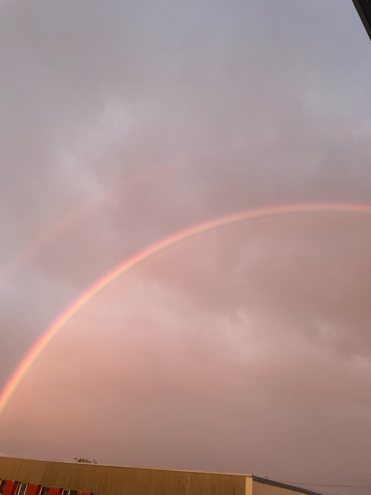 The image depicts a double rainbow arcing across a cloudy, overcast sky, tinged with pink and gray hues, possibly indicating a sunset or sunrise. The sky fills the majority of the frame. Below the sky, a portion of a building is visible at the bottom, featuring a light brown facade with some red elements along the lower edge. The building appears to be a low-rise structure. In the distance, silhouetted trees can be seen against the sky. The overall scene suggests an outdoor environment, possibly in a semi-urban or suburban area. The image is a photograph taken in Tartu, Estonia.