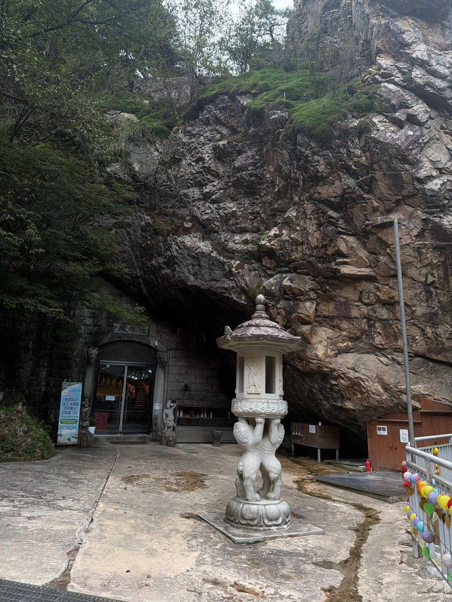 The image shows the entrance to a cave temple, likely located in the mountainous region surrounding Dongducheon-si, South Korea. The entrance is a large opening in a rocky cliff face, partially covered by vegetation. In front of the entrance stands a stone lantern, intricately carved and supported by figures of two cherubic beings. The pathway leading to the entrance is paved with concrete, showing a prominent crack. To the left of the entrance, a blue informational sign is posted. To the right, a wooden structure and a fire extinguisher are visible, along with a metal railing adorned with colorful paper lanterns. The overall impression is one of a serene, natural setting integrated with religious architecture.