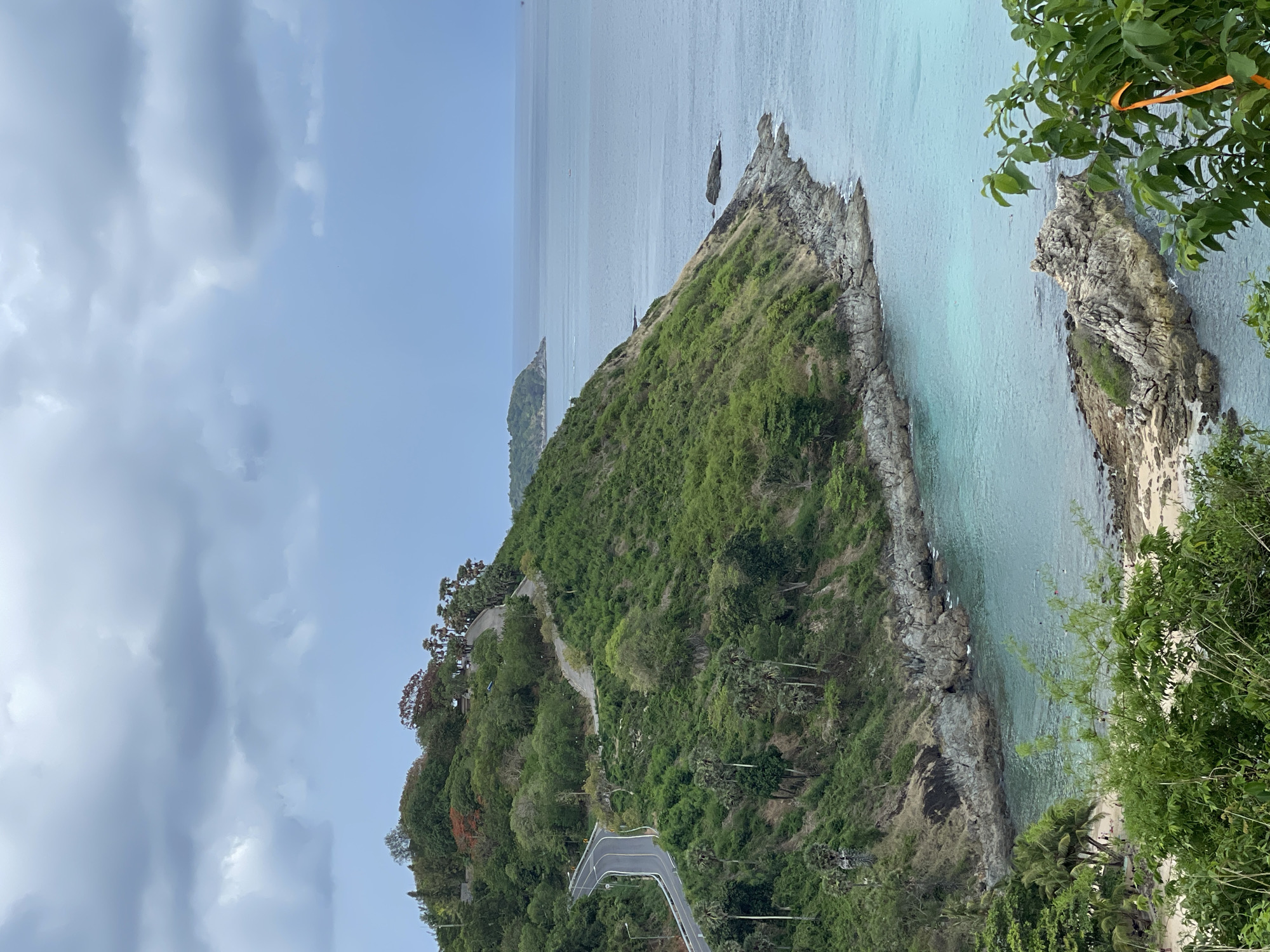 The image depicts Promthep Cape, a scenic viewpoint located in Rawai, Thailand. The viewpoint is characterized by a grassy, rocky promontory jutting into the turquoise sea. Lush green vegetation covers the cape, with a winding road visible. A smaller rocky islet is seen adjacent to the main cape, surrounded by water. The sky is overcast with a mix of blue and gray clouds. A glimpse of another landmass can be seen in the distance.