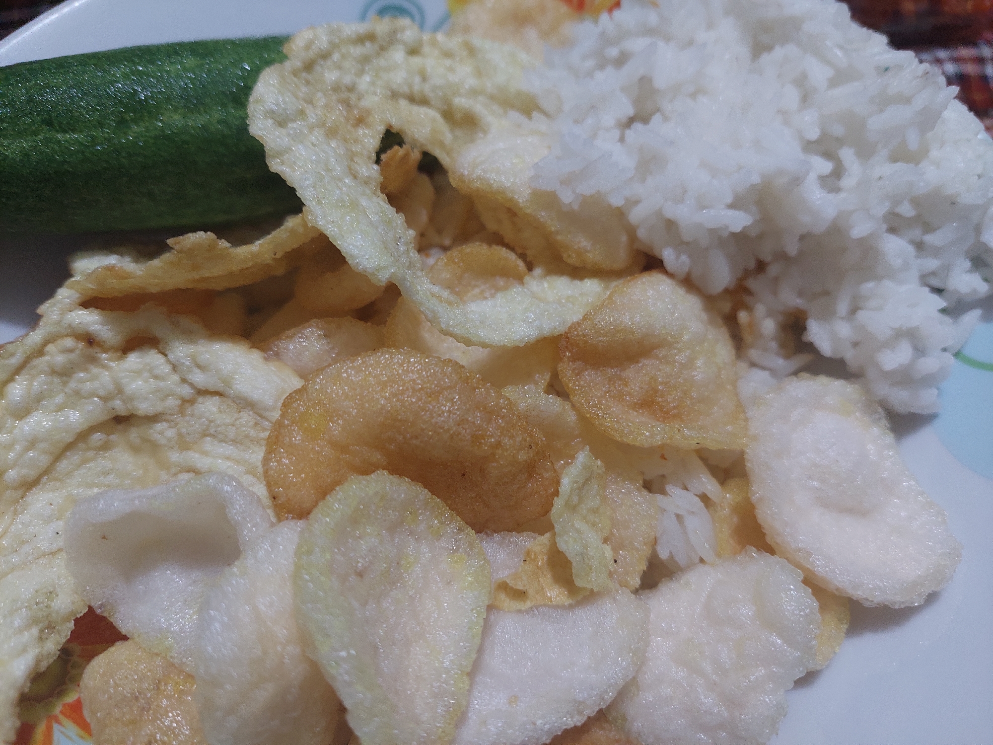 A close-up, overhead shot shows a plate of food. On the left and center of the plate are white and golden-brown puffed crackers, layered over white rice. A piece of green cucumber is visible on the right side of the plate. The food appears to be a light meal, possibly breakfast or a snack, and it is served on a white plate with a colorful pattern around the rim. The setting is likely a home or casual dining establishment in Jelutung, Indonesia. No individuals are visible in the image.