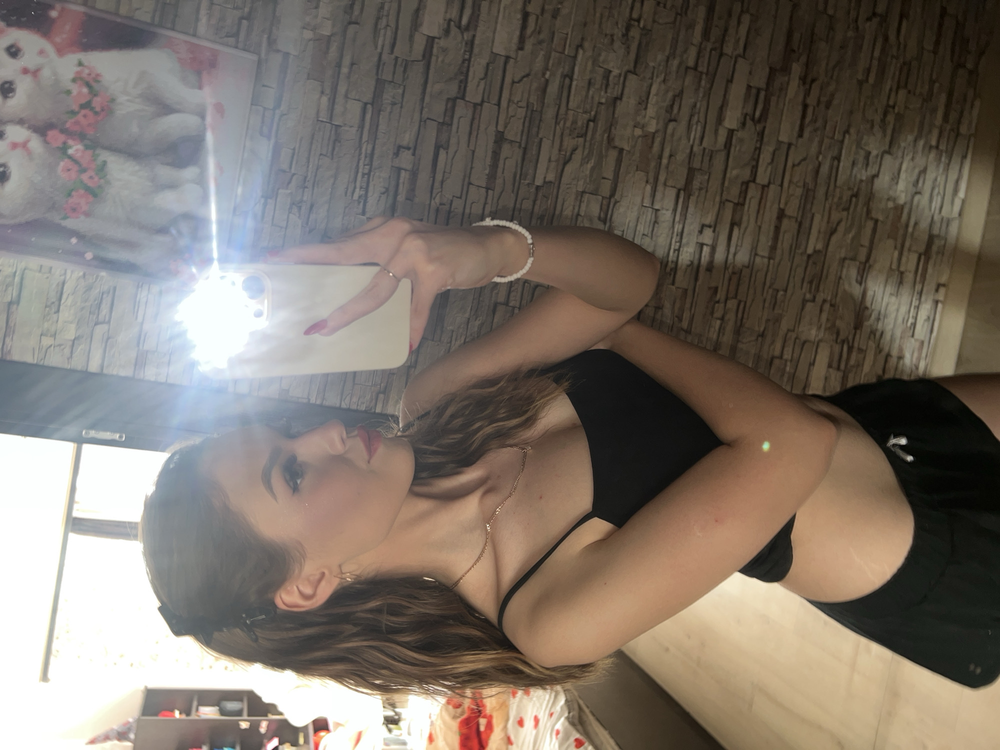 A young woman with brown hair pulled into a ponytail is taking a mirror selfie in a room. She is wearing a black tank top and black shorts. She has on a thin gold necklace and a white beaded bracelet. Behind her, a picture of two white cats wearing floral collars is visible on a stone-patterned wall. To her left, a window with blinds is visible, and a shelf with items is partially seen.
