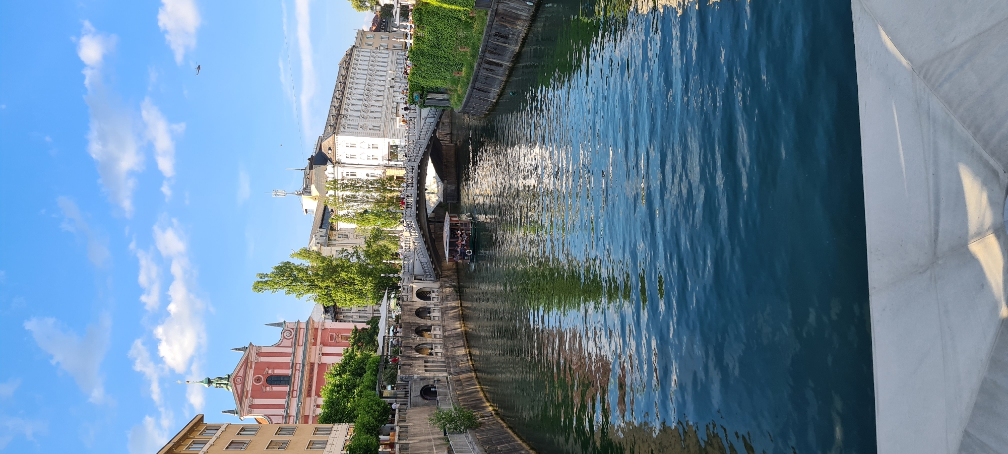 The image depicts a scenic view of the Ljubljanica River in Ljubljana, Slovenia, under a clear blue sky with scattered white clouds. In the foreground, the dark blue water of the river fills the lower half of the frame, reflecting the surrounding architecture and greenery. A white, modern-looking structure, possibly a railing or part of a bridge, occupies the very bottom of the image.
A tour boat with several passengers is navigating the river, moving from left to right. The boat is dark green with a brown roof.
On the left bank of the river, a light-colored building with arched walkways is visible, with trees providing shade. Further back, a pink and white church with a green-domed steeple dominates the skyline, with other buildings and lush green trees behind it.
On the right bank, a grand white building with numerous windows and ornate architectural details stands prominently. A white pedestrian bridge, adorned with railings and people walking across it, spans the river, connecting the two banks. The right bank is also lined with a wall covered in vibrant green ivy, and more trees are visible in the background.
In the upper right portion of the sky, a small dark object, possibly a plane or drone, is visible. The overall scene conveys a peaceful urban atmosphere with historical and natural elements.