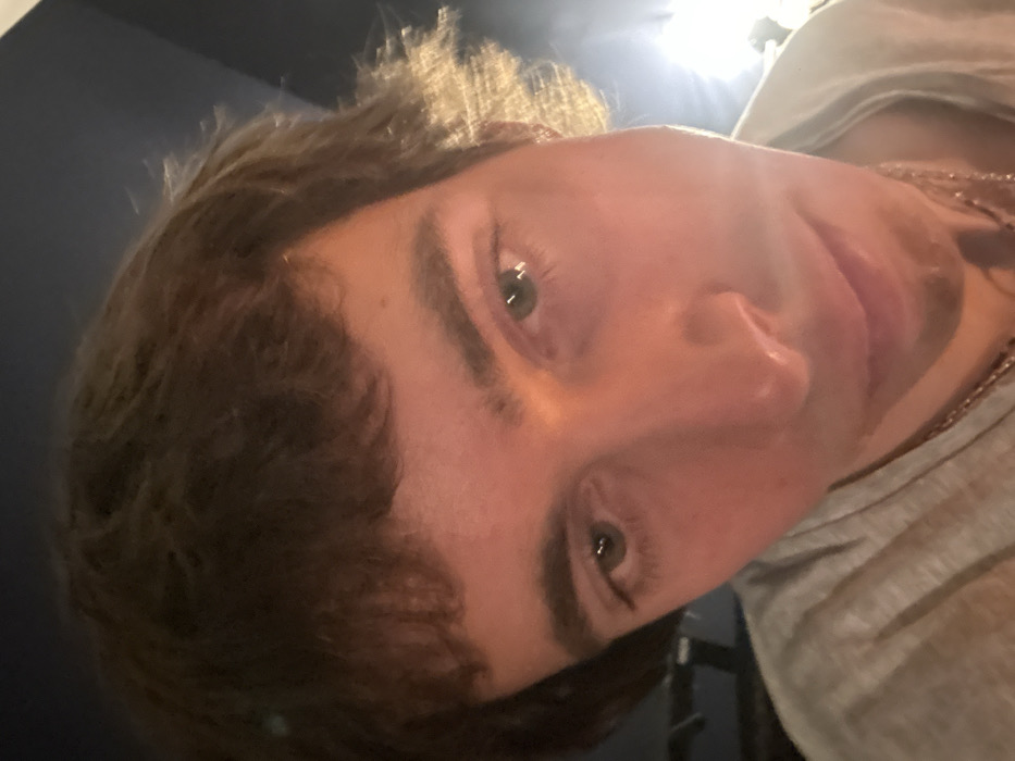 A close-up, slightly low-angle photograph features a young adult male with fair skin, light brown, shaggy hair with bangs, prominent eyebrows, and green/hazel eyes, looking towards the camera with a neutral expression. He is wearing a light-colored t-shirt and a visible chain necklace. The background is dark and out of focus, with a bright, artificial light source causing a flare on the right side, illuminating the hair on that side. Another light source appears to illuminate the subject's face, casting a subtle reddish tint. The specific location within Paris, France, is not identifiable from the visual content of the image.