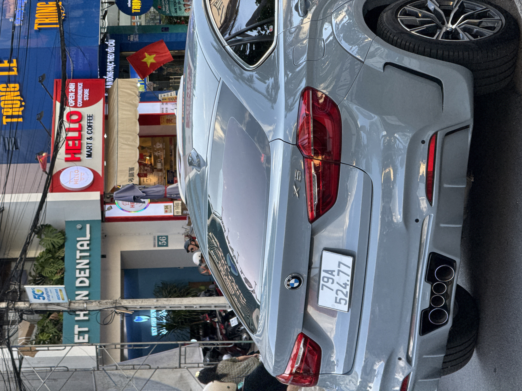 A light gray BMW X6 with a Vietnamese license plate (79A 524.77) is parked on a city street. The car's rear is prominently displayed, showing its quad exhaust pipes and the BMW logo. In the background, a street scene unfolds with various shops, including "VIET HAN DENTAL" and a "HELLO MART & COFFEE" store. A Vietnamese flag is visible, hanging from a pole. People are seen on the sidewalk, some wearing helmets, suggesting the presence of motorcycles, a common mode of transport in Vietnam. The environment appears to be an urban setting in Nha Trang, Vietnam, characterized by buildings, overhead wires, and signage in Vietnamese.