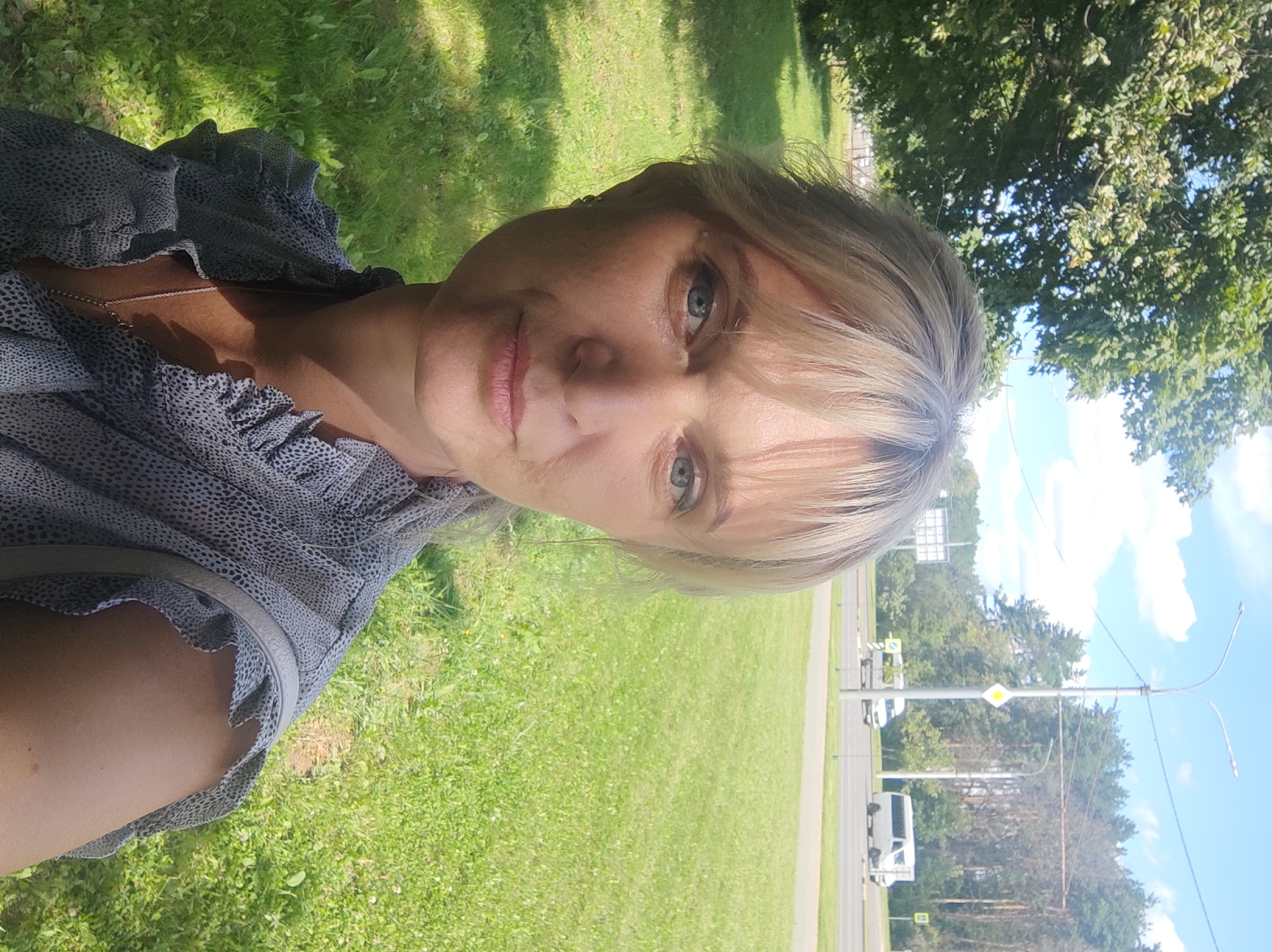The image is a selfie taken outdoors on a sunny day, likely in Mahilyow, Belarus. The subject is a woman with blonde hair and blue eyes, wearing a gray patterned blouse. She is standing in a grassy area with trees and a road visible in the background. Two white vans are driving on the road, and streetlights and traffic signs are also present. The overall scene suggests a casual outdoor setting, possibly a park or a roadside in a suburban or urban area.