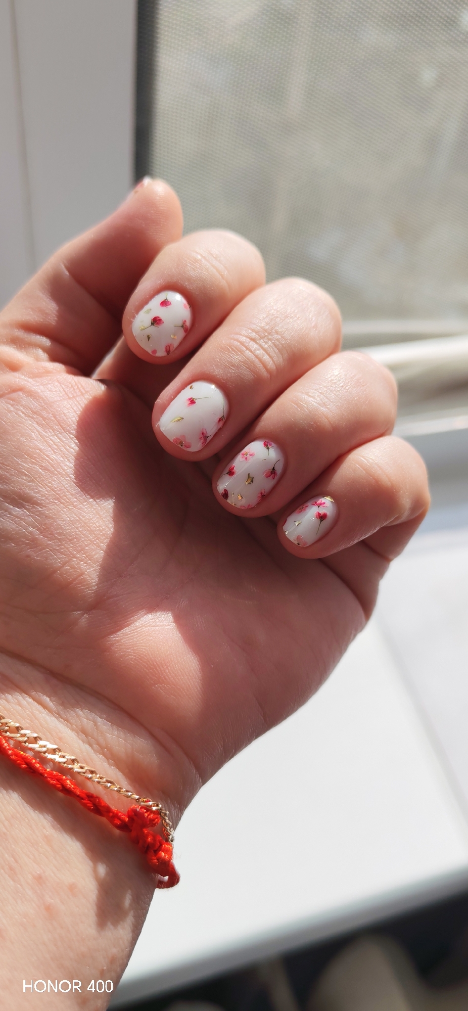 A close-up photograph shows a person's left hand against a blurred background of a window and possibly a white sill. The nails are short and rounded, painted with a white base coat and decorated with small pink floral patterns and gold foil accents. The hand is slightly clenched, with fingers bent. A red and gold bracelet is visible on the wrist. The lower left corner of the image contains a "HONOR 400" watermark, suggesting the device used to capture the image.
