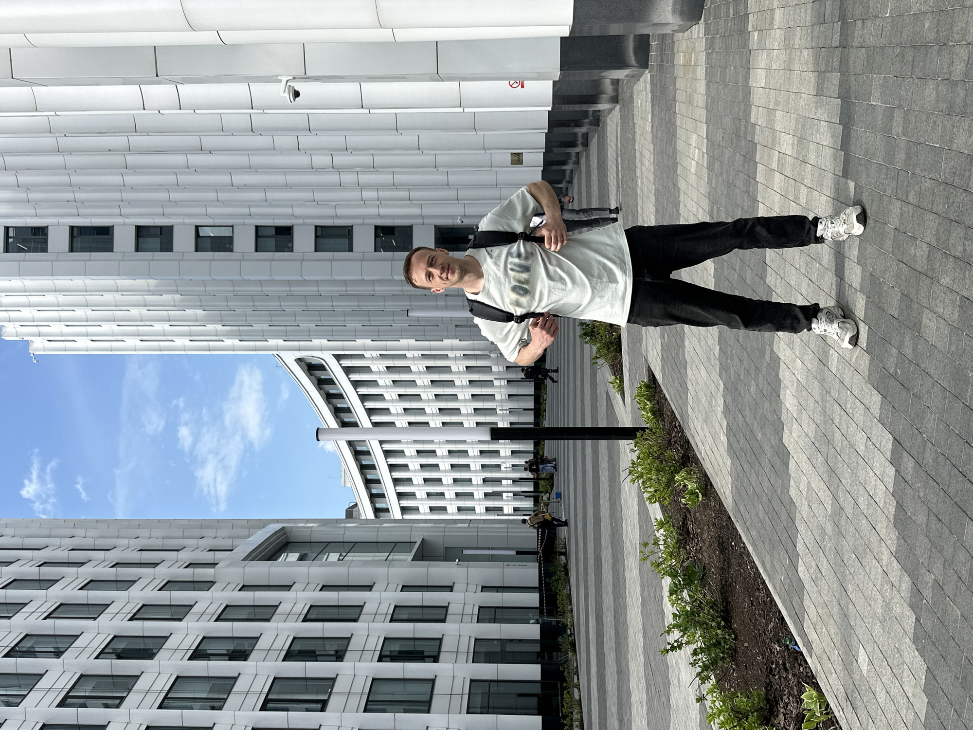 The image shows a man with short brown hair standing on a grey brick walkway in Moscow, Russia. He is wearing a light grey t-shirt with writing on it, black pants, white sneakers, and a black backpack. The man is looking directly at the camera with a slight smile. To the left and behind the man are several modern buildings with a large number of windows. There is a section of blue sky visible between the buildings. A tall black pole is next to the walkway. On the right side of the walkway are plants growing in a narrow dirt bed. Several people can be seen walking in the distance behind the man.