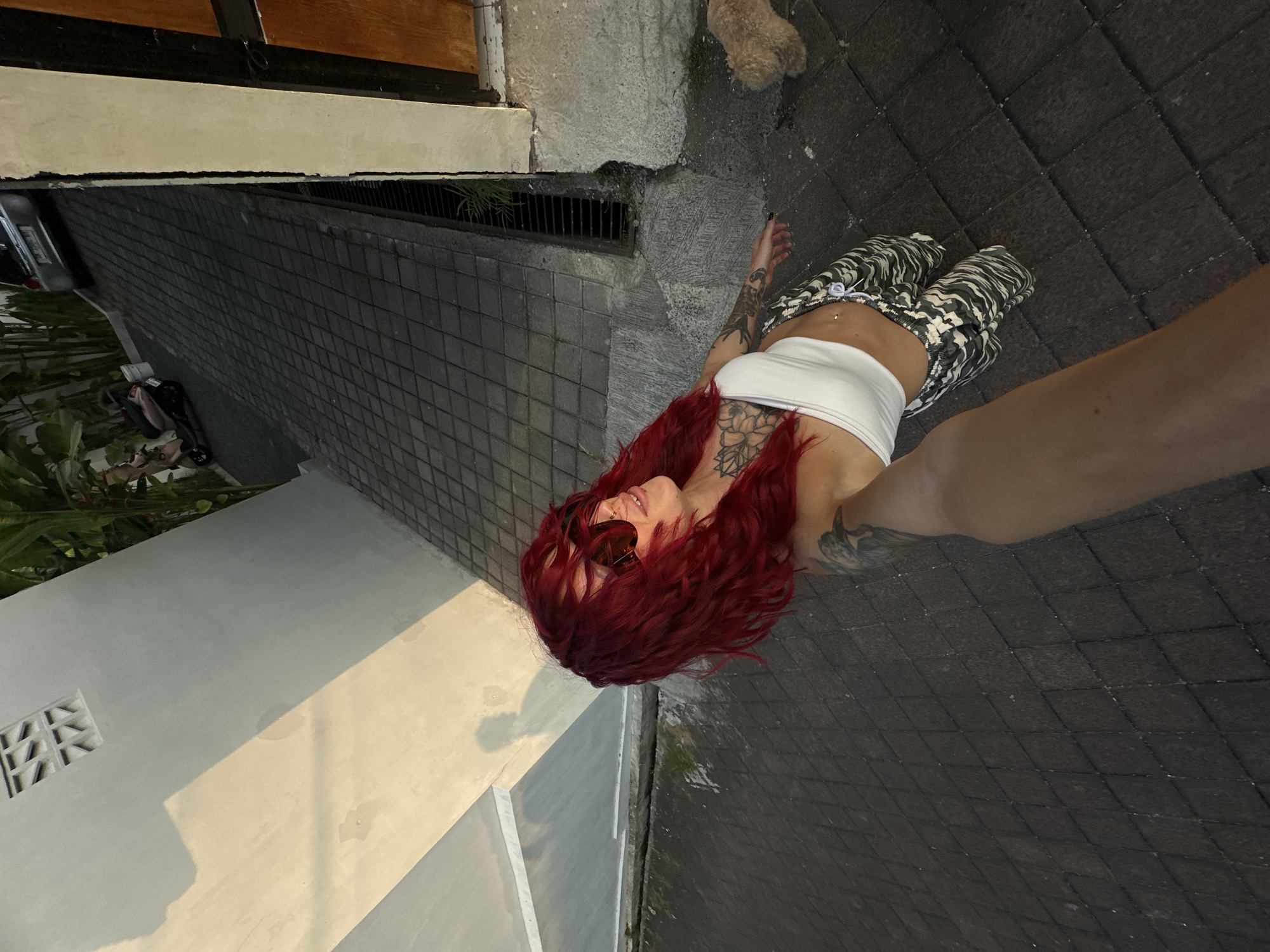 A woman with vibrant red, curly hair and sunglasses is posing outdoors. She is wearing a white crop top and camouflage-patterned pants. She has visible tattoos on her chest and arm. The background shows a paved pathway leading uphill, with a white building to the left and a wooden door to the right. A small, fluffy dog is visible near the door. A scooter and a car are parked further up the pathway. The lighting suggests it is either sunrise or sunset. The scene appears to be located in North Kuta, Indonesia, possibly a residential area or a pathway leading to a property.