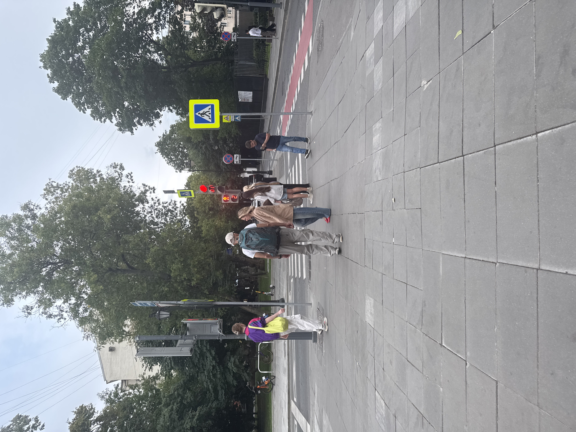 A pedestrian crossing on a cloudy day in Moscow, Russia. A group of pedestrians, including individuals wearing backpacks, are gathered at the crossing, some waiting for traffic lights to change while others are in motion. Traffic signals, pedestrian crossing signs, and street lamps are visible. The street is paved with asphalt, and the sidewalk is made of gray tiles. Trees line the background, with buildings and fences visible in the distance.