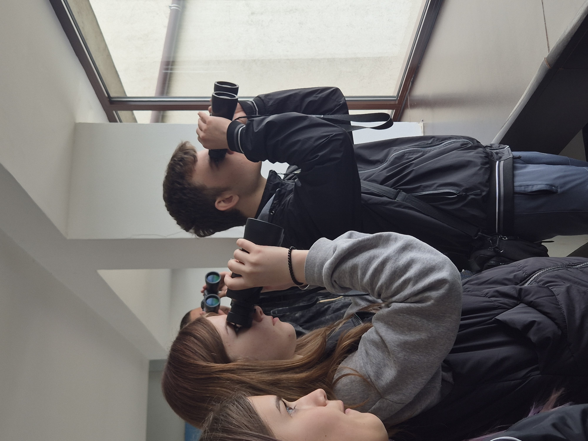 Two young women and a young man stand indoors in Srebarna, Bulgaria, looking upward through binoculars. The man is wearing a black jacket and has short brown hair. One woman is wearing a gray sweatshirt and has reddish-brown hair, and the other has dark brown hair. They appear to be looking out of a window.