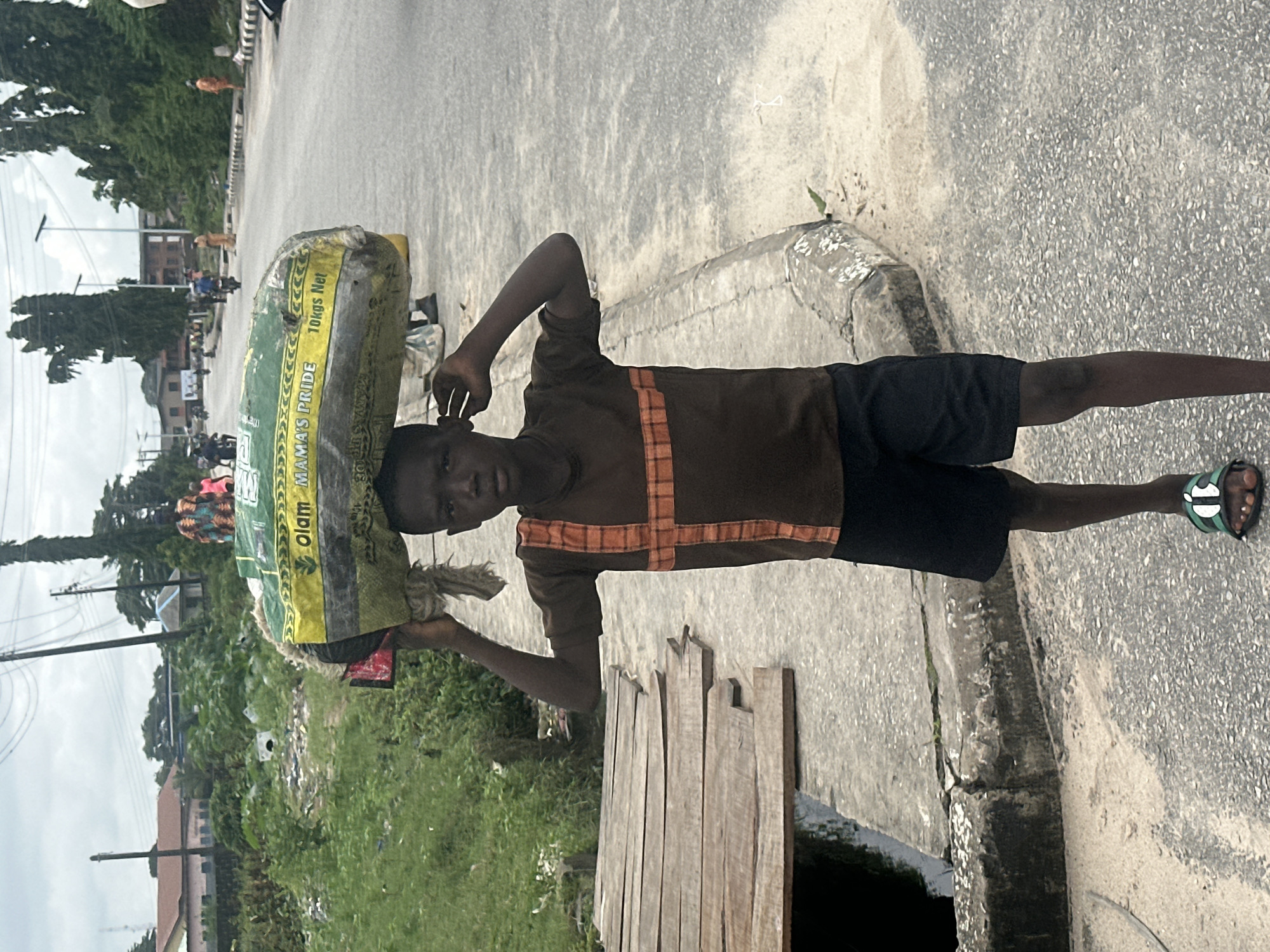A young boy, possibly of African descent, is captured in a medium shot, standing on a paved road in Badagry, Nigeria. He is wearing a brown t-shirt with an orange plaid design across the chest and black shorts. On his head, he balances a large, green and yellow sack labeled "OLAM MAMA'S PRIDE 10Kgs Net". His right hand is raised to steady the load, while his left hand is touching his ear. He wears black flip-flops with a white and green design. The boy stands near a concrete curb and a makeshift wooden bridge crossing a dark, water-filled channel. In the background, a street stretches into the distance, lined with trees and buildings. Other people and vehicles are visible further down the road, suggesting a bustling urban environment. The sky is overcast, indicating a cloudy day.