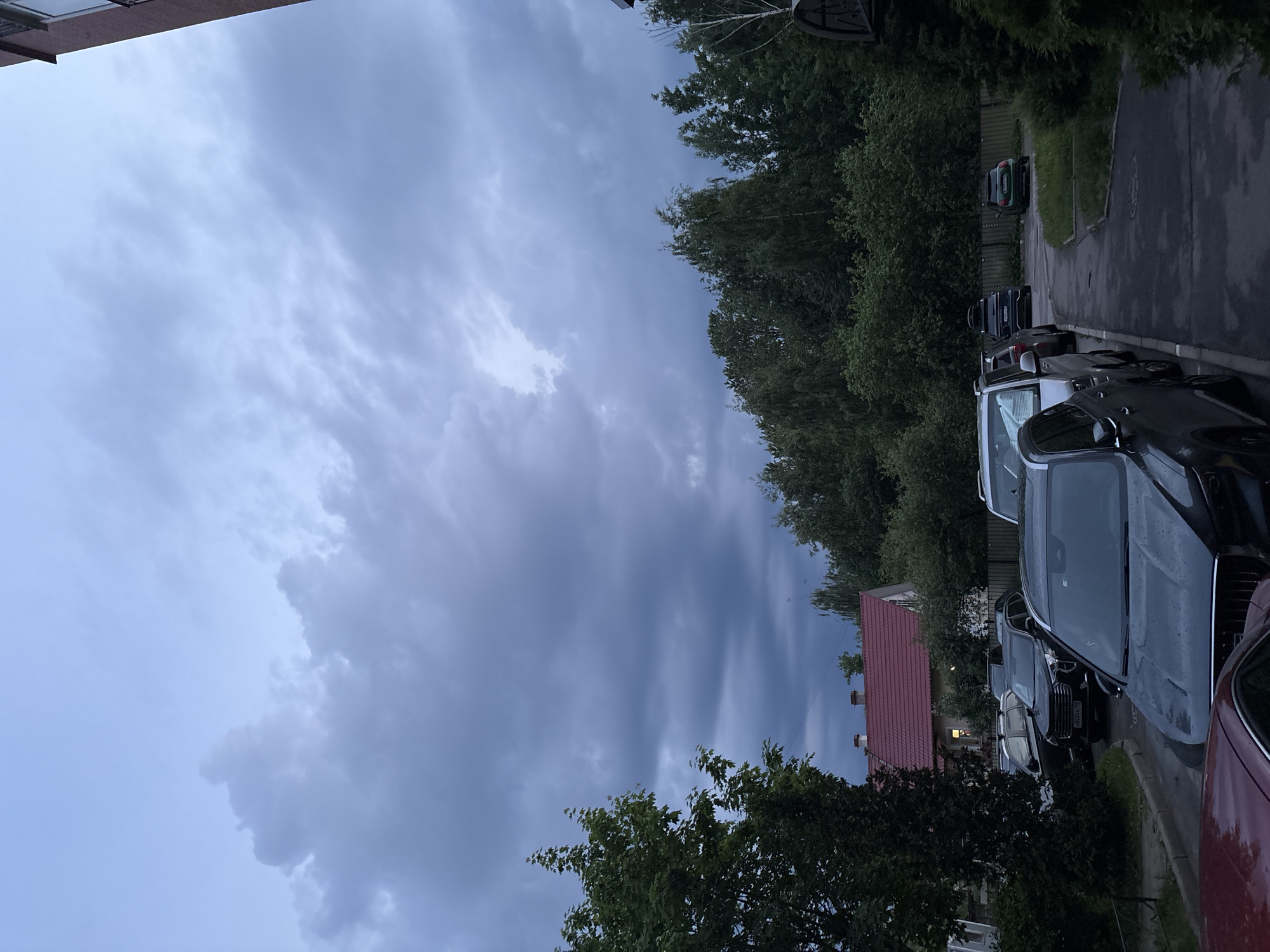 The image depicts a residential street scene in Saint Petersburg, Russia, under a heavily overcast, dark grey sky at twilight or early evening. Dense, undulating clouds dominate the upper two-thirds of the frame, with a slightly lighter blue-grey tone at the very top. The ground, including the asphalt path and parking areas, appears damp, possibly from recent rain.
In the mid-ground, multiple automobiles are parked along the side of the path. A dark-colored SUV, possibly a Haval, is prominently visible with Russian license plate "H043XA 198", confirming the location in Saint Petersburg. Other parked vehicles, including several SUVs and a red car partially visible in the extreme foreground, are present.
To the left, a large, leafy deciduous tree with dark green foliage partially obscures the view of a multi-story house or building featuring a distinct red-tiled roof and light-colored walls. A single window in this building glows with a warm, interior light. Further background is characterized by a dense treeline of various deciduous trees, with a wooden fence running along their base, separating them from the parking area.
On the far right, the partial facade of a taller, light-brown building with visible windows frames the scene, suggesting a multi-story residential or commercial structure. No people are visible in the image. The overall atmosphere is quiet and subdued, indicative of a damp, overcast evening in a green residential district.