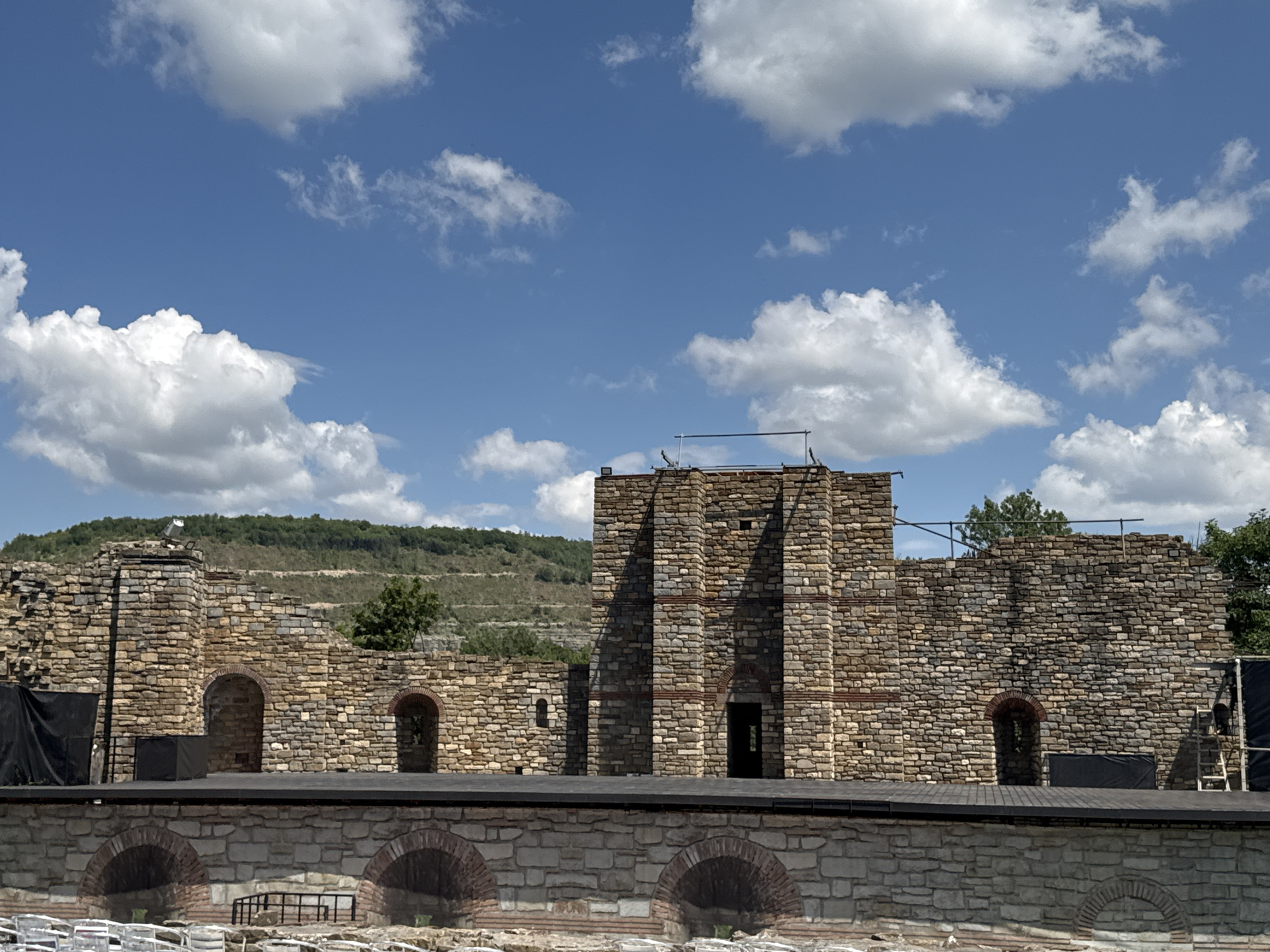 This is a medium-angle, eye-level, outdoor, daytime shot of the Ancient Theatre of Philippopolis, located in Plovdiv, Bulgaria, which is often mistakenly associated with Veliko Tarnovo. The ancient stone amphitheater, partially reconstructed and repurposed as a performance venue, dominates the center of the frame. The weathered stone walls, constructed from irregular blocks of various earthy tones, display arched openings and tiered seating sections that extend towards the background. A dark wooden stage platform is set in front of the lower section of the amphitheater, with black fabric panels and a ladder visible on the right side. In the foreground, rows of white plastic chairs are arranged, suggesting an audience seating area. The background features a lush, green hillside dotted with trees, and the sky above is a vibrant blue, adorned with scattered white cumulus clouds. The overall scene evokes a sense of historical preservation and contemporary use of an ancient site.