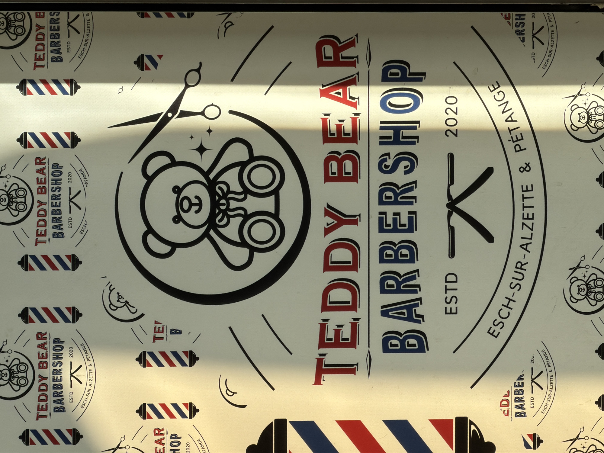 The image shows a promotional banner or sign for "Teddy Bear Barbershop," located in Esch-sur-Alzette, Luxembourg. The main text "TEDDY BEAR" is in red, and "BARBERSHOP" is in blue. A teddy bear logo is centrally placed, surrounded by a circle, and paired with barber-related graphics such as scissors and barber poles. The text "ESTD 2020 ESCH-SUR-ALZETTE & PÉTANGE" is incorporated into the design, indicating the barbershop's founding year and geographical area. The background pattern consists of smaller teddy bear logos and barber poles. The lighting across the image suggests a nearby window or light source, casting shadows across the surface.