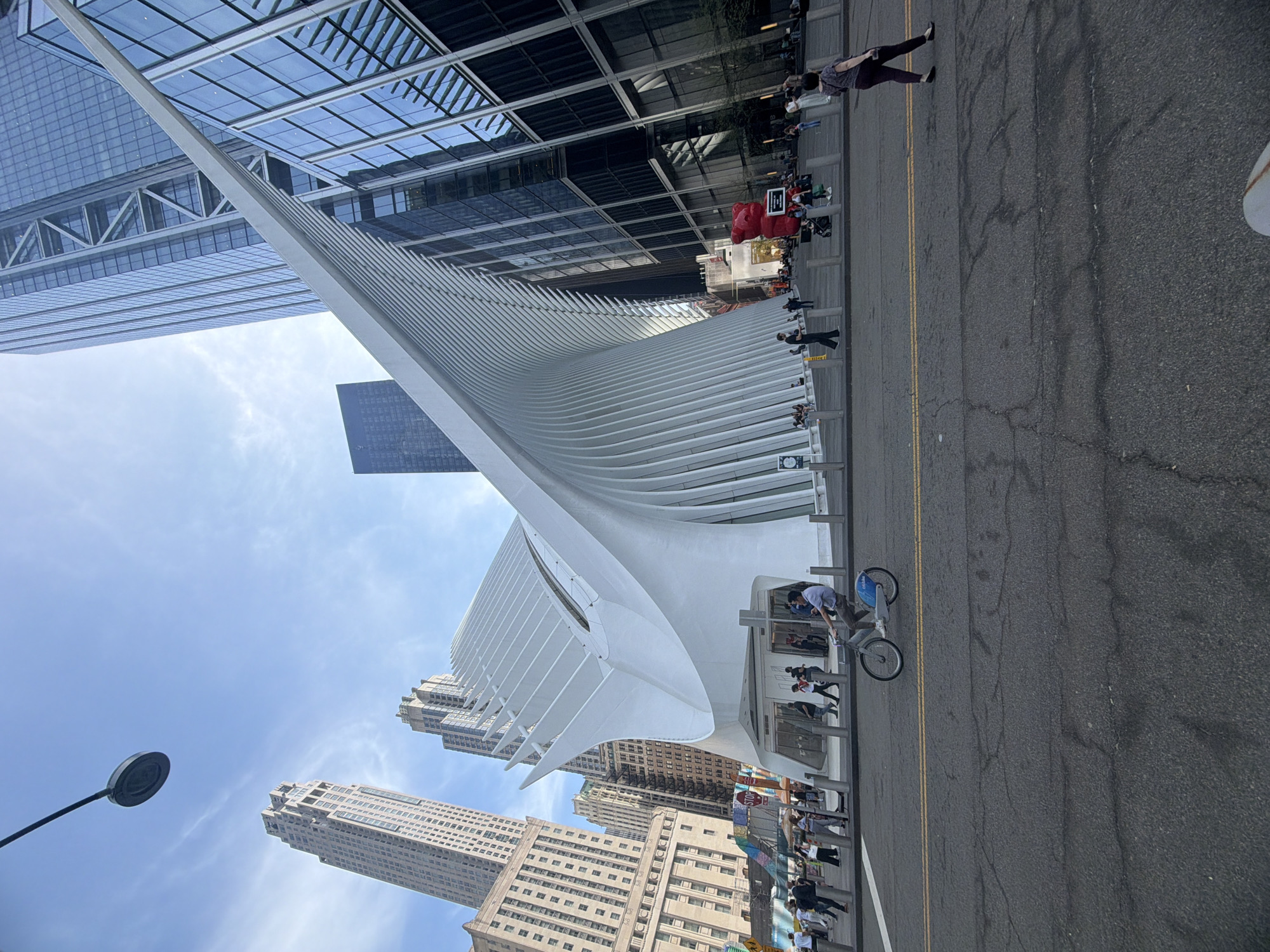 The media file captures a vibrant urban scene in New York City, dominated by the striking, white architectural form of the World Trade Center Transportation Hub, known as the Oculus. The image is taken from street level, looking up at the surrounding skyscrapers and the intricate design of the hub.
The Oculus, with its distinctive white, ribbed structure, stretches across the left and central parts of the frame, appearing to open up towards the sky. Behind and around it, numerous tall, modern buildings with reflective glass facades rise prominently against a clear, bright blue sky, some showing reflections of the clouds and other structures. A darker, rectangular skyscraper stands further back in the distance.
On the right side of the image, an asphalt street or paved area is visible, marked by distinct yellow double lines. Several people are seen on the sidewalk and along the street. One individual is captured riding a blue and white bicycle, positioned near a "Citibike" docking station which clearly displays the "Citibike" logo. Other pedestrians are walking, standing, or gathered in small groups near the base of the buildings and along what appears to be a public walkway with planters. In the mid-ground, a large, bright red object, possibly a sculpture or inflatable structure, is visible near some people.
Notable details include a street lamp in the lower-left corner and a partially visible "STOP" sign on a pole further down the street. The bright lighting and clear sky indicate a sunny daytime setting, likely late morning or afternoon. The scene conveys a sense of a busy, modern metropolis with architectural grandeur and daily urban activity.