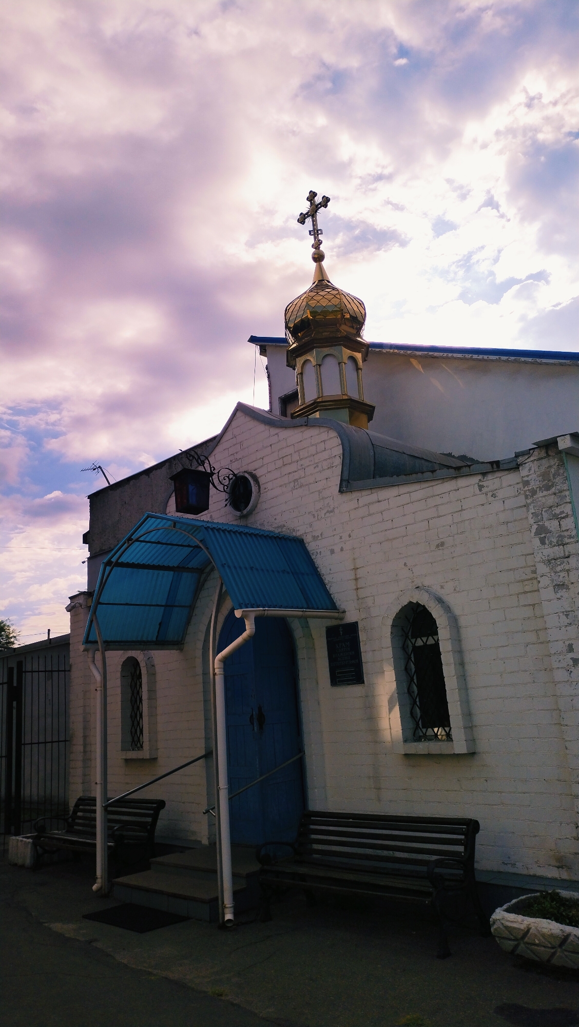 The image shows a white brick building with a golden dome topped with a cross, characteristic of an Orthodox church. The building has a blue door and a blue awning over the entrance. There are two dark wooden benches in front of the building, one on each side of the entrance steps. A fence is visible to the left of the building. The sky is cloudy with hues of pink and purple, suggesting it might be dawn or dusk. The architecture and the presence of an Orthodox cross indicate this is likely a religious building, possibly a church or chapel. The general appearance of the building and its surroundings suggests an urban or semi-urban setting in Ukraine. Specifically, this building is the Holy Transfiguration Church in the city of Horishni Plavni, Ukraine. There are no people visible in the image.