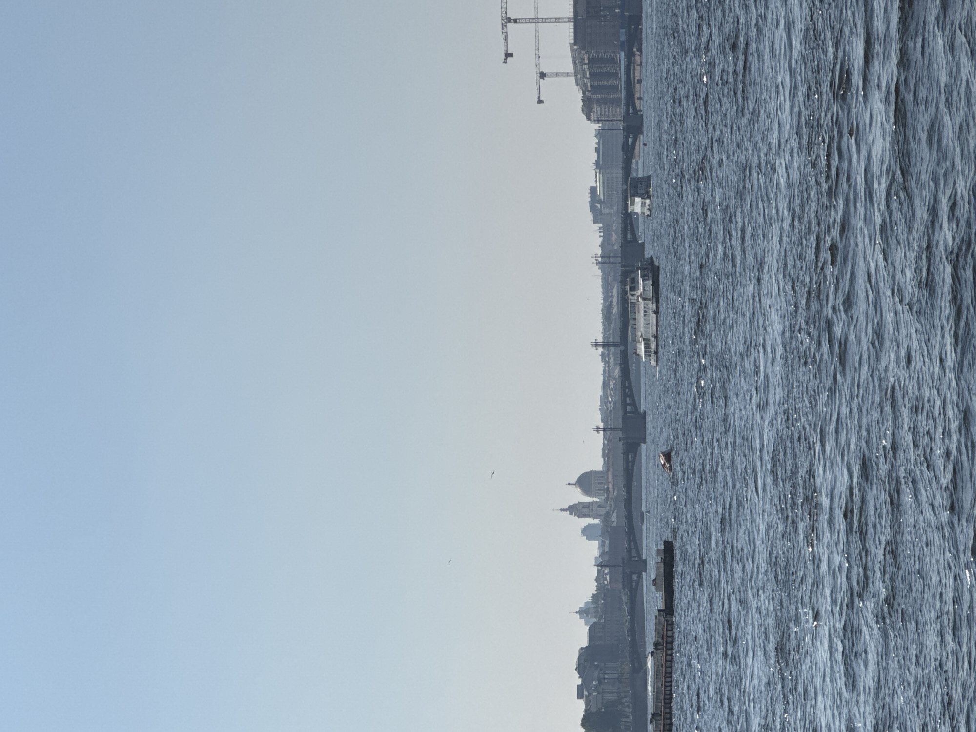 The image captures a wide view of the Neva River in Saint Petersburg, Russia. The river is characterized by its choppy, textured surface, reflecting the bright sky. On the right side of the frame, the water dominates, showing its dynamic movement. The left side of the image is a vast expanse of clear, pale blue sky, suggesting a bright, possibly early morning or late afternoon.
Along the riverbank, a city skyline is visible, featuring a mix of modern and historical architecture. Prominent structures include cranes, indicating ongoing construction or development. There are also several buildings with domed roofs, a characteristic feature of Saint Petersburg's architectural style, such as the dome of Saint Isaac's Cathedral, which is discernible in the distance. Several boats and ferries are present on the water, suggesting active river traffic. The overall atmosphere is serene, with the gentle movement of the water and the distant city creating a sense of urban tranquility. The clear sky implies fair weather. No text is visible in the image.