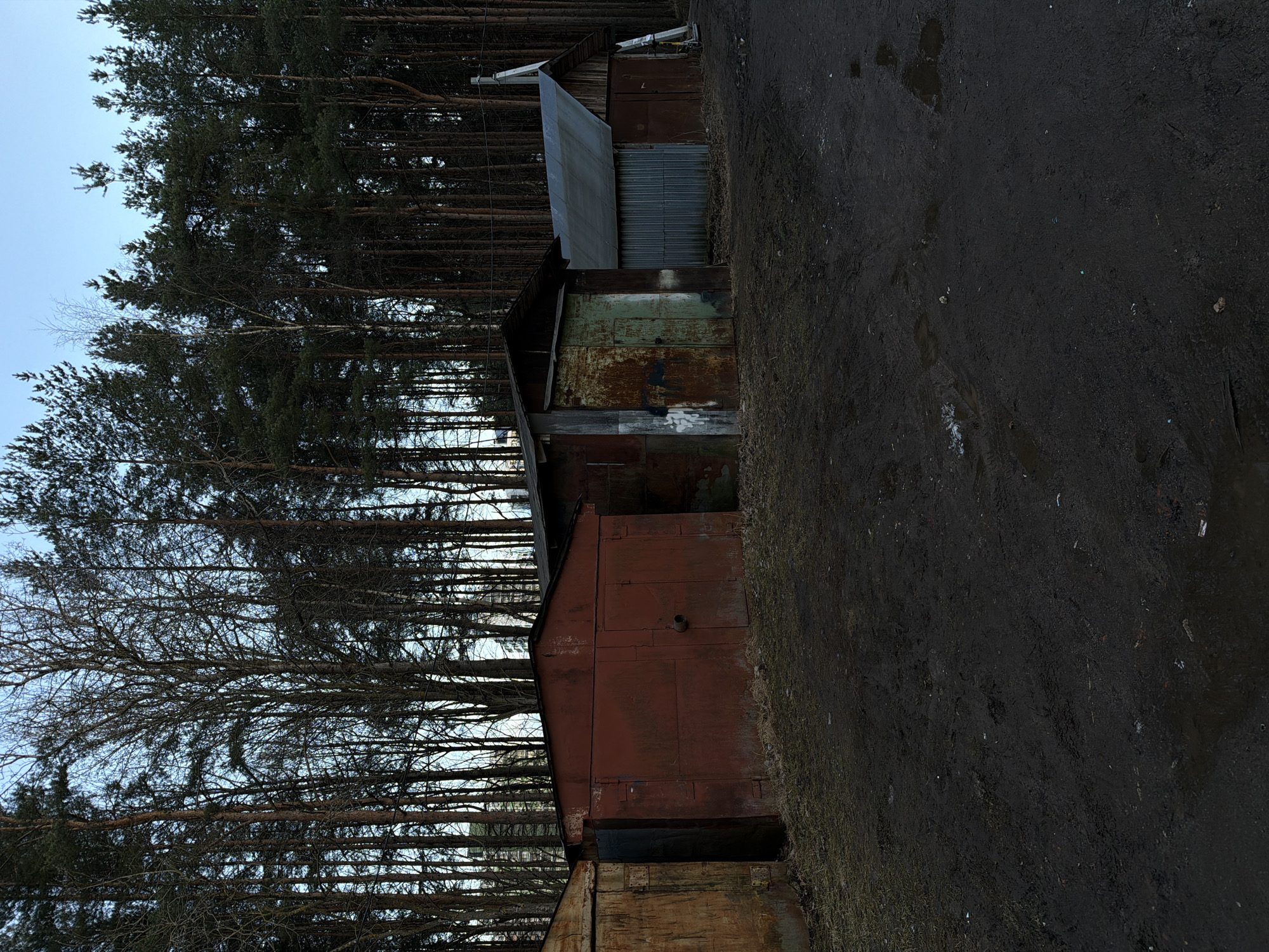 The image depicts a row of derelict garages situated in Lavriki, Russia. The garages are constructed from corrugated metal and wood, exhibiting signs of rust, decay, and weathering. A dense forest of tall, bare trees forms the backdrop. The ground in the foreground appears to be a dirt path or roadway, muddy and wet. The sky is overcast, suggesting a grey and damp day. There are no people visible in the scene.