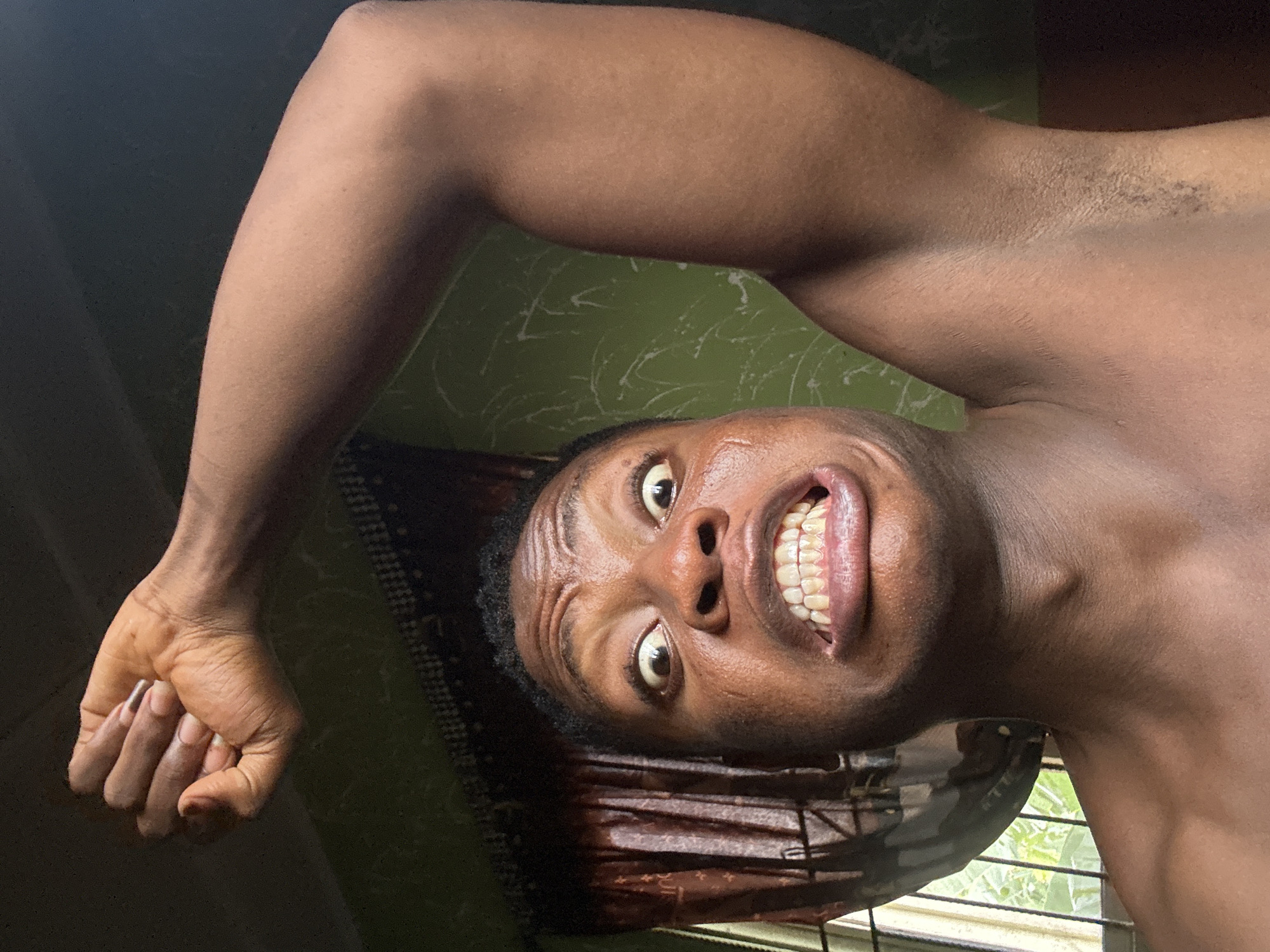 The image shows a close-up, angled shot of a shirtless individual, likely a young adult male with dark skin, photographed from a low perspective. The person is looking directly at the camera with an intense, wide-eyed expression and an open mouth that reveals prominent, white teeth, resembling a grimace, exaggerated smile, or a look of surprise/exertion. Beads of sweat are visible on their forehead and cheeks, suggesting warmth or recent activity.
One arm is raised and bent, showcasing a muscular bicep, while the hand is clenched into a fist with visible fingernails. Short, dark hair is present on the head and in the armpit area. The background features a solid green wall or curtain behind the upper body, and a window with brown patterned blinds or curtains below the chin, allowing natural light to illuminate the scene. Green foliage is visible outside the window, confirming it is daytime. The scene is set in Ikorodu, Nigeria. There is no visible text in the image.