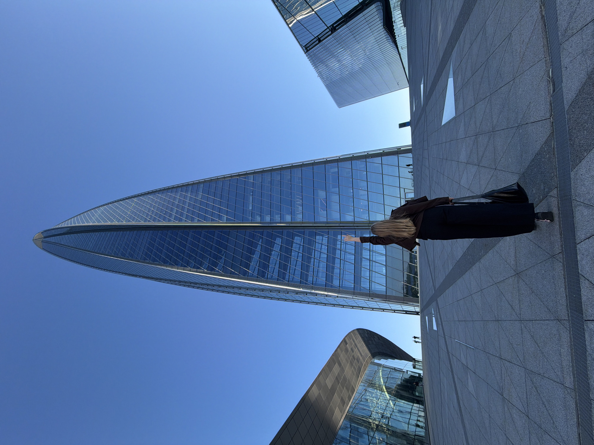 In the city of Saint Petersburg, Russia, a blonde-haired woman with long hair, wearing a brown jacket and black pants, stands on a wide, gray, tiled plaza, facing away from the camera. Her right arm is raised with her index finger pointing upwards. She carries a black handbag in her left hand. The plaza is located in front of a modern architectural complex featuring a very tall, slender, glass skyscraper with a pointed top, and two lower, angular, glass-faced buildings on either side. The sky is clear and blue.