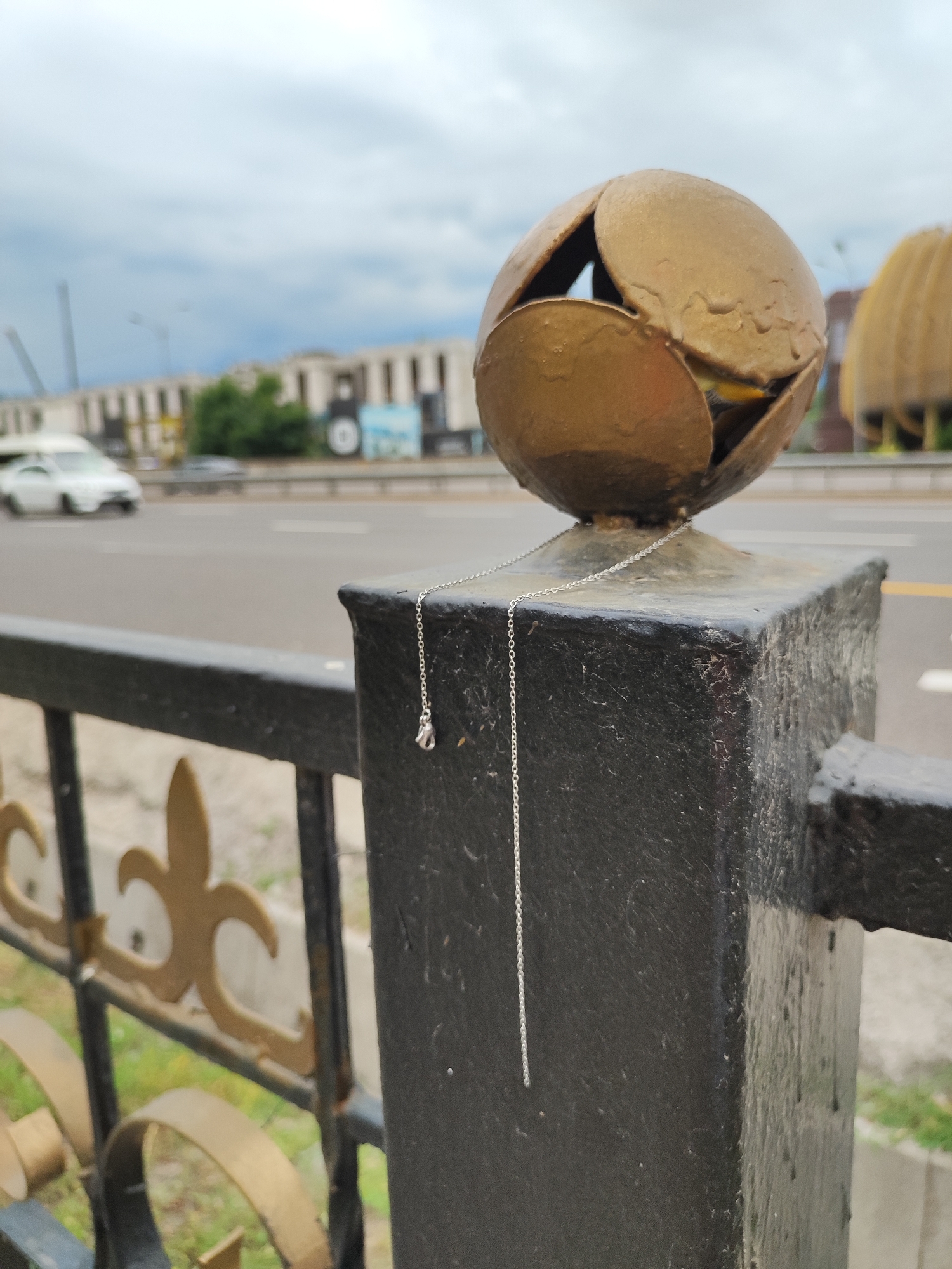 A silver necklace with a small diamond pendant drapes over a black metal post topped with a golden, petal-shaped sculpture. The post is part of a black metal railing with decorative golden fleur-de-lis motifs. Behind the railing, a multi-lane road is visible with blurred vehicles, indicating it is likely a street or highway in a city. In the background, a long, light-colored building with a repetitive facade suggests an urban setting. The sky is overcast with grey clouds. The scene appears to be captured in Almaty, Kazakhstan, given the distinctive golden sculptures that are common architectural elements in the city, particularly in public spaces and along avenues like Dostyk Avenue.