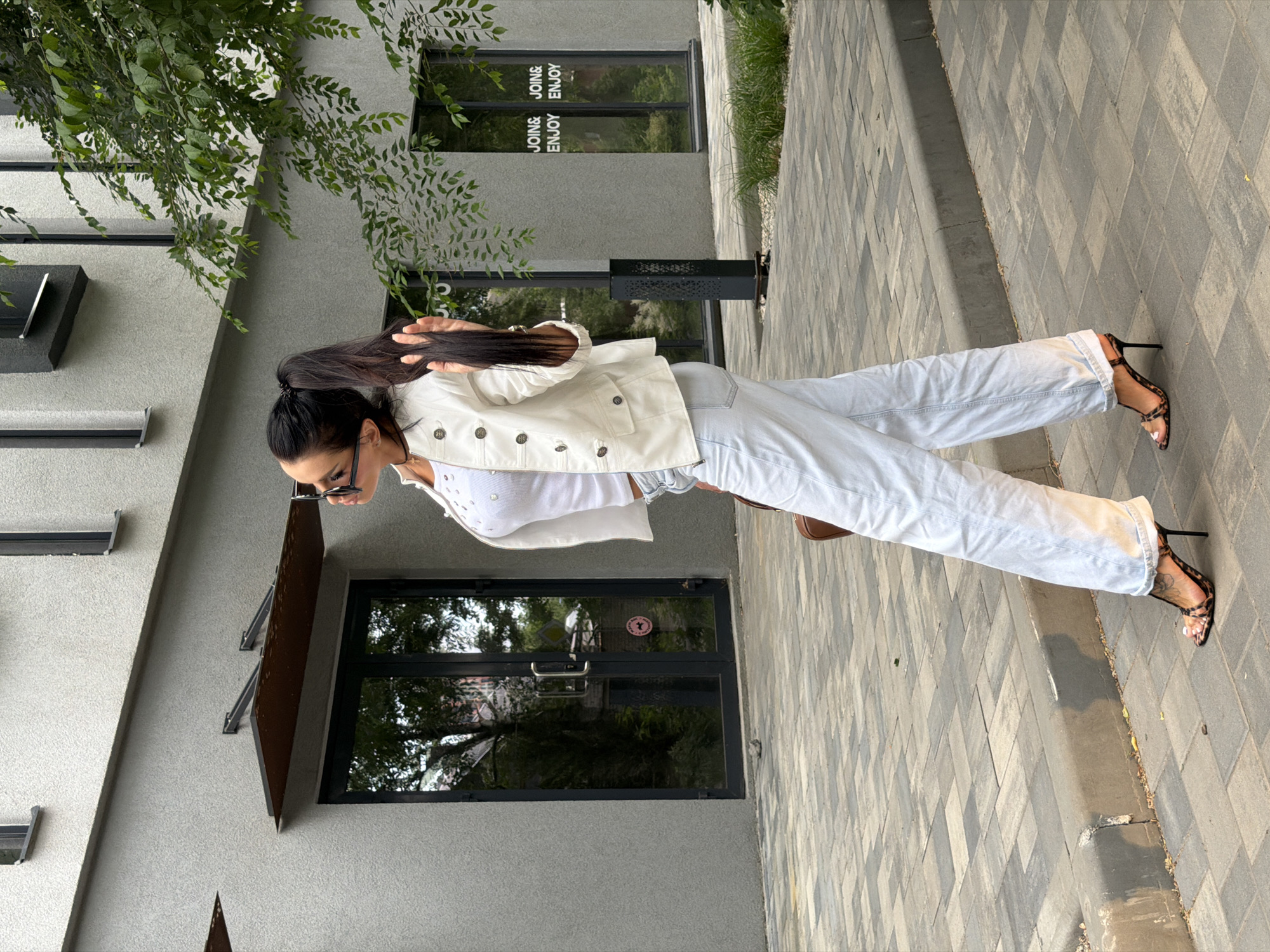 A person stands on a paved walkway in front of a modern building. The person is wearing a white denim jacket over a white top and light-colored jeans. They have on strappy high-heeled sandals with a leopard print pattern. Their long, dark hair is pulled back into a ponytail. They are looking down and to the side, and appear to be adjusting their sunglasses.
The building has a grey textured facade with dark-framed windows and a dark awning over a doorway. Green foliage is visible in the upper left corner, suggesting it's an outdoor setting. The walkway is made of grey rectangular pavers.
Reflections in the window show trees and an outdoor scene. Text is visible on the windows of the building, reading "JOIN& ENJOY" repeated. The lighting suggests it is daytime, possibly late afternoon due to the angle of the shadows and the warm light. The overall impression is casual and stylish.
