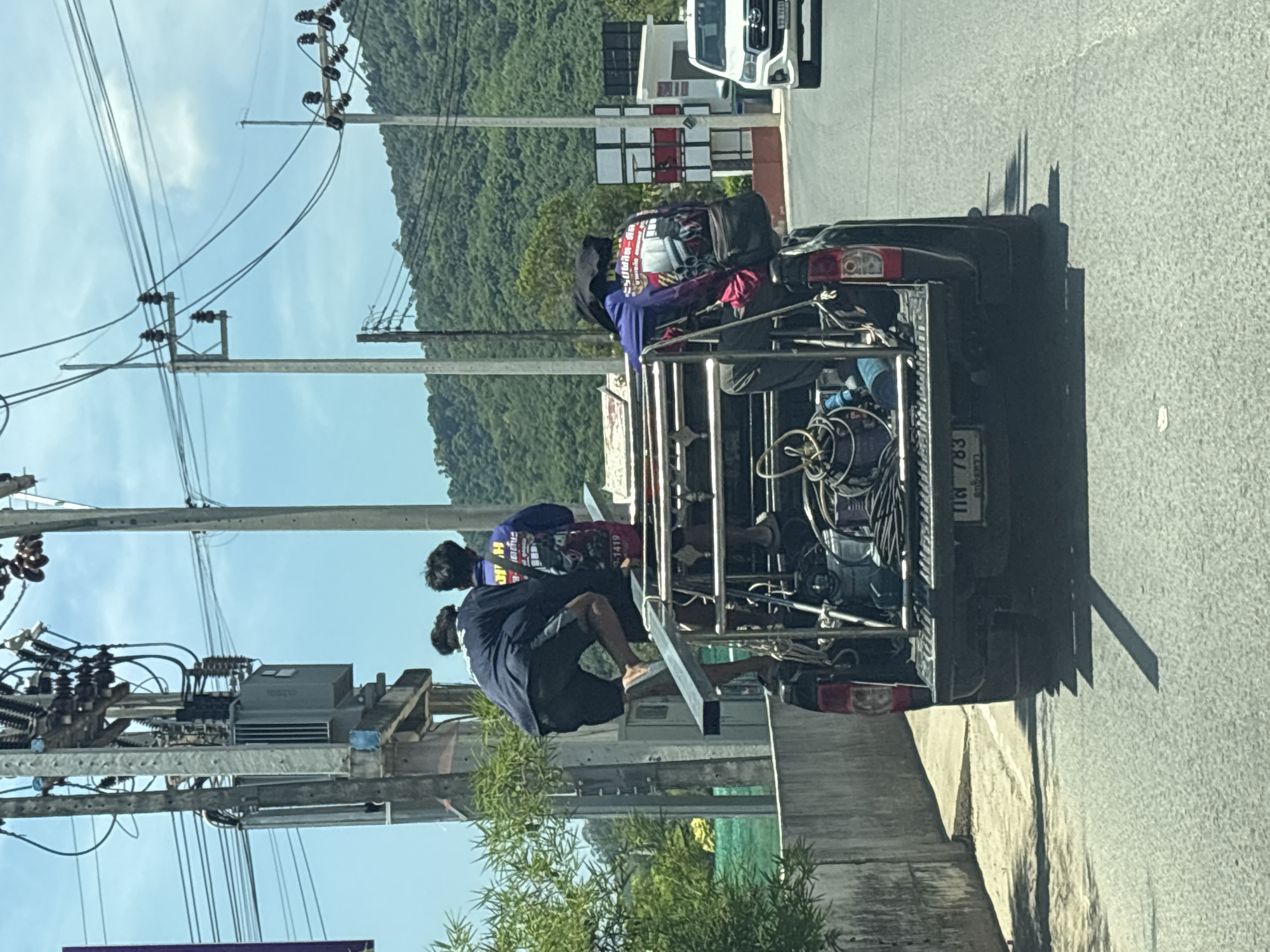 The image shows a black pickup truck traveling on a road in what appears to be Choeng Thale, Thailand, as evidenced by the Thai license plate "tin 783". Three people are riding in the truck bed, which has a metal frame structure. The bed is filled with equipment, including coiled cables and other items. The background features a blue sky, mountains, and power lines. A white car is visible further up the road. Vegetation is present in the foreground.