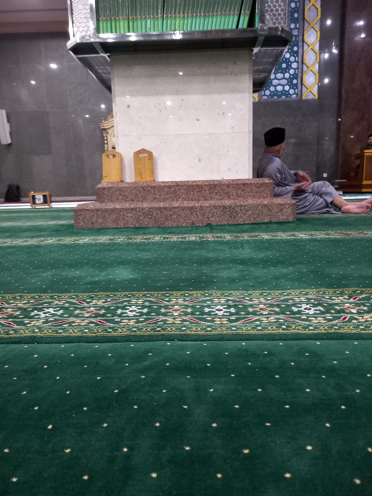 The image depicts the interior of a mosque in Samarinda, Indonesia. A man, dressed in a traditional gray peci and robe, sits on a raised step covered in speckled brown carpet. His legs are crossed, and his hands are clasped. The mosque features a large, green carpet with intricate patterns in gold, red, and white. In the background, a shelf holds a row of green-covered books, likely Qurans. Two wooden stands with cut-out handles are visible near the steps. The walls are a mix of dark grey and marble-like finishes, with decorative blue and yellow tilework on the right side. Natural and artificial lights illuminate the space.