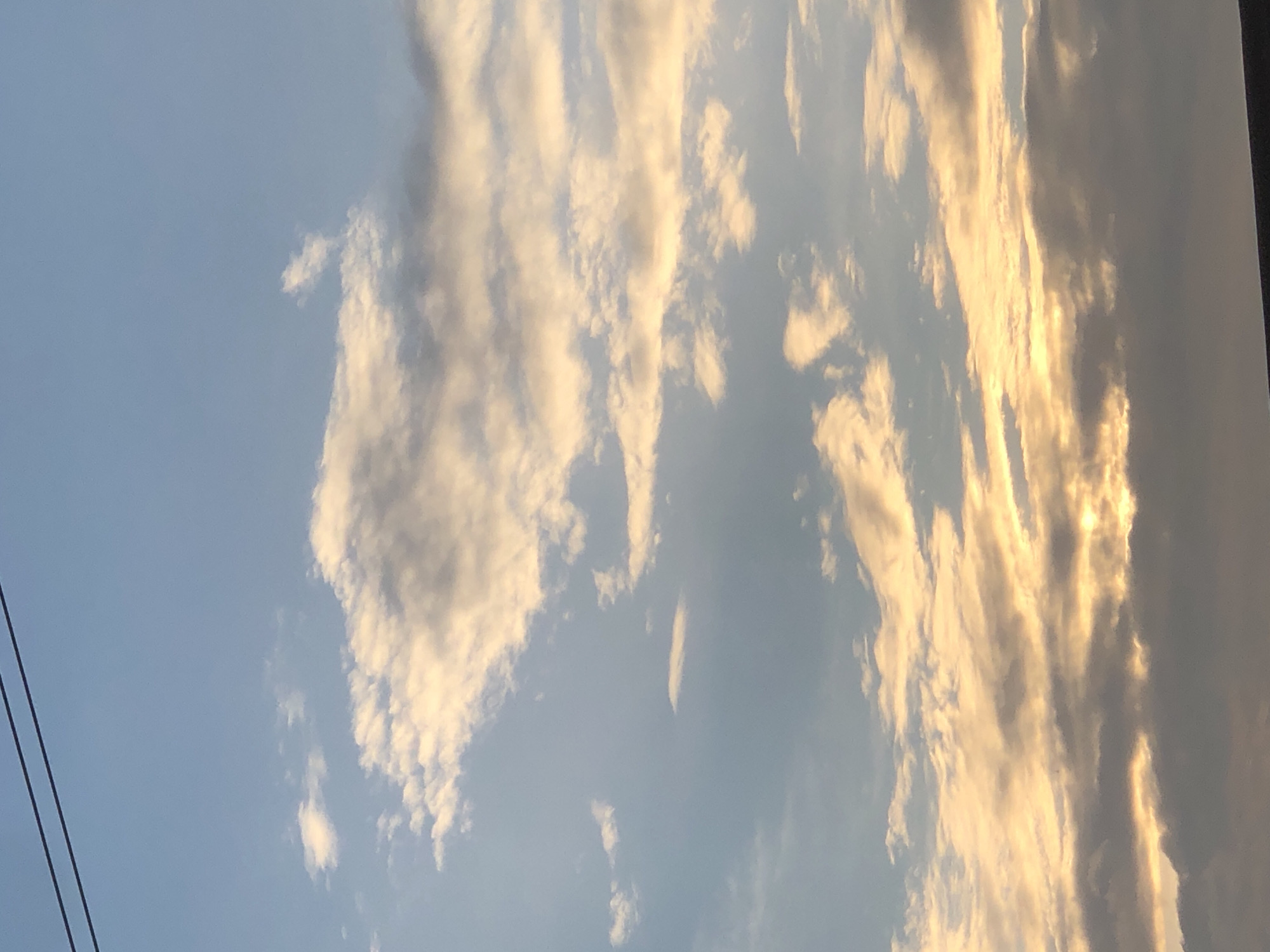 The image shows a sky with clouds at dusk, possibly in Ajah, Nigeria. Two dark power lines are visible in the upper left corner of the frame. The clouds are illuminated by the setting sun, casting a warm, golden glow on some of them, while others appear darker and more shadowed. The sky transitions from a pale blue at the top to a warmer hue near the horizon, where the sun is likely setting, though it is not directly visible. The overall impression is one of a serene evening sky.