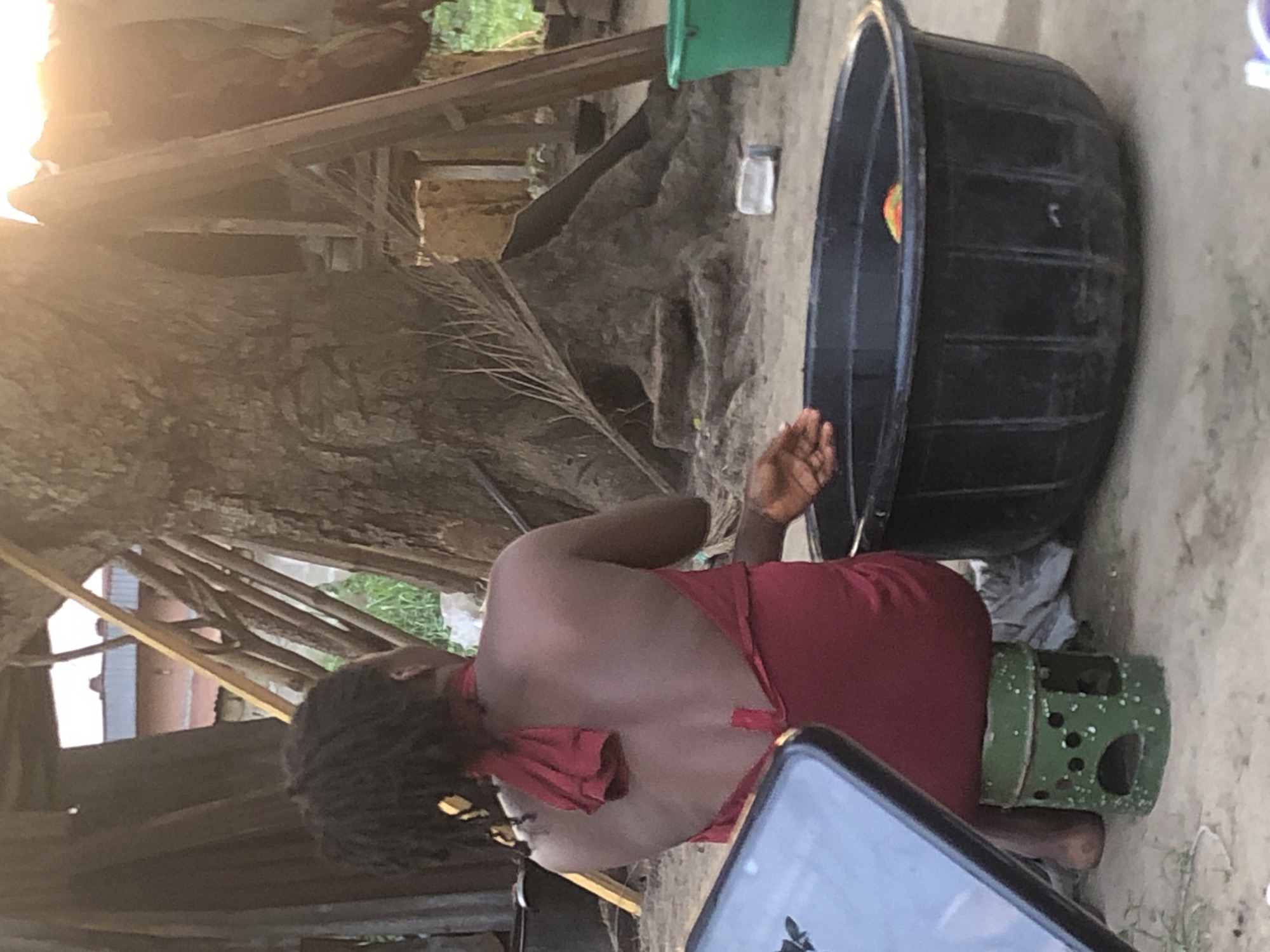 A woman with dreadlocks, wearing a red dress with a low back, is crouching next to a large black tub outside in Atani, Nigeria. She is seated on a green perforated container. A green bucket and a small white container are nearby. The scene is under a partly constructed or dilapidated wooden structure. The ground is light colored. A smartphone partially obstructs the view in the foreground.
