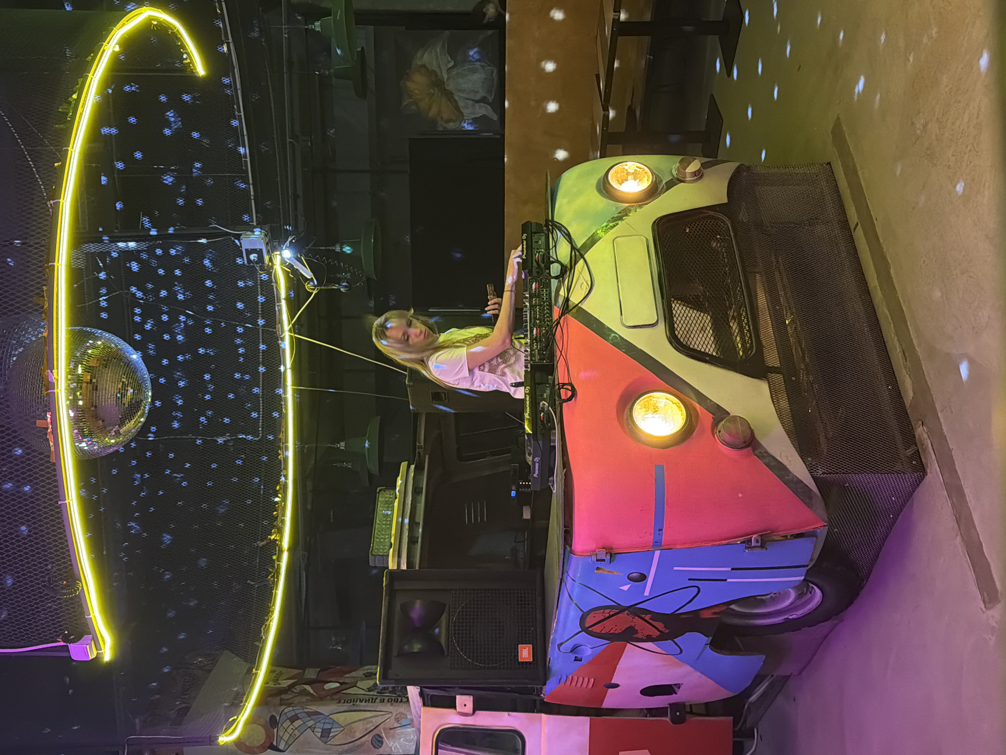 An active indoor nightlife scene is depicted in Saint Petersburg, Russia, centered around a female DJ performing. A woman with blonde hair, wearing a light-colored t-shirt, stands behind a distinct DJ setup, operating a Pioneer DJ mixer/controller. She holds a smartphone in her left hand, appearing focused on her task.
The DJ booth is creatively constructed from the front section of a vehicle, possibly a vintage van or bus, featuring two illuminated circular headlights and a mesh grille. Its exterior is painted with a vibrant, multi-colored abstract design in segments of red, blue, green, and white, including a stylized abstract symbol on the blue part. A JBL speaker, with its brand name visible, is positioned to the left of the booth.
The venue's atmosphere is accentuated by striking lighting. A prominent, curved yellow neon light fixture arches over the left side of the scene. A disco ball is also visible, casting numerous small, scattered blue and white light reflections onto the dark background and the light-colored floor, creating a lively, party-like ambiance. The setting suggests an evening event with a dynamic visual and auditory experience.