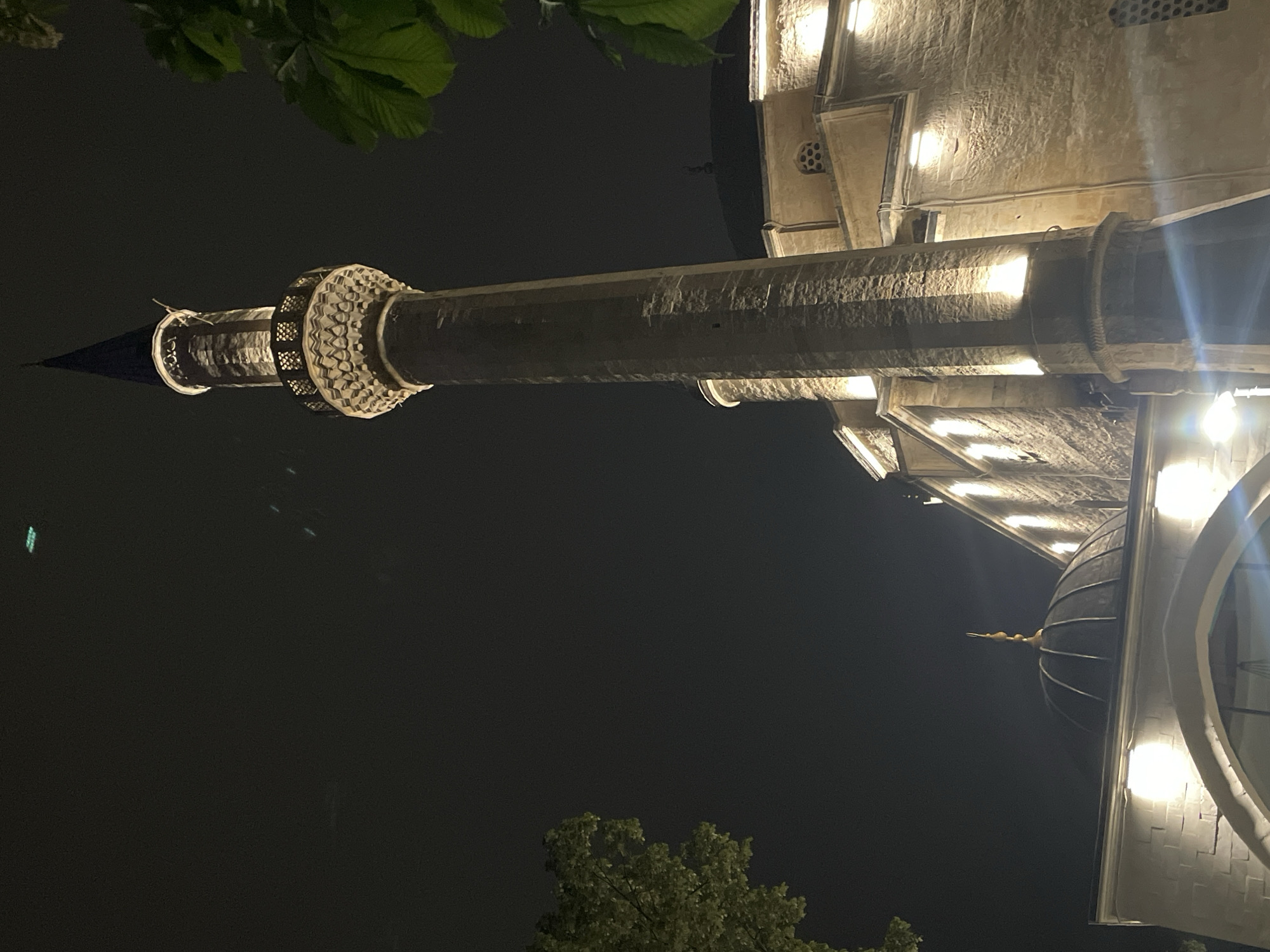 The image shows a minaret of a mosque at night, illuminated by artificial lights. The mosque appears to be constructed of light-colored stone or concrete. The minaret is a tall, cylindrical structure topped with a conical roof. The sky is dark, and some tree branches are visible at the top and bottom of the image. The location is Razgrad, Bulgaria.
