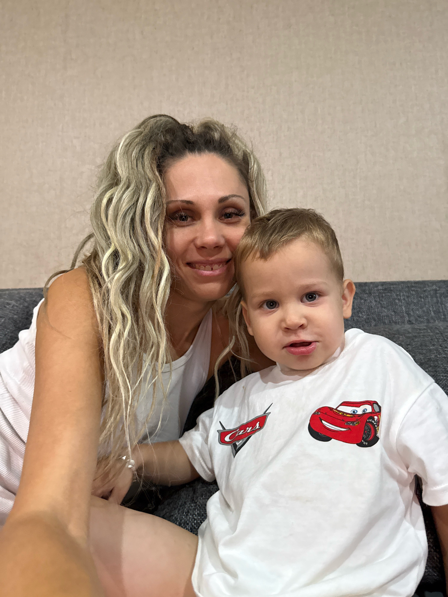 This media file is a selfie of a woman and a young boy, likely mother and son, sitting on a couch in an indoor setting. The woman has long, wavy blonde hair with highlights, and is wearing a white tank top. She is smiling and looking at the camera. The boy, with light brown hair and blue eyes, is wearing a white t-shirt featuring characters from the Disney movie "Cars." He is also looking towards the camera with a curious expression. The background consists of a textured, neutral-colored wall and the dark fabric of the couch. The lighting suggests it is daytime, but no specific time of day can be determined. There is no indication of the weather or precise location beyond the general context of Rusanovo, Russia. The overall emotion conveyed is one of warmth and familial connection. The t-shirt has visible text which reads "Cars".