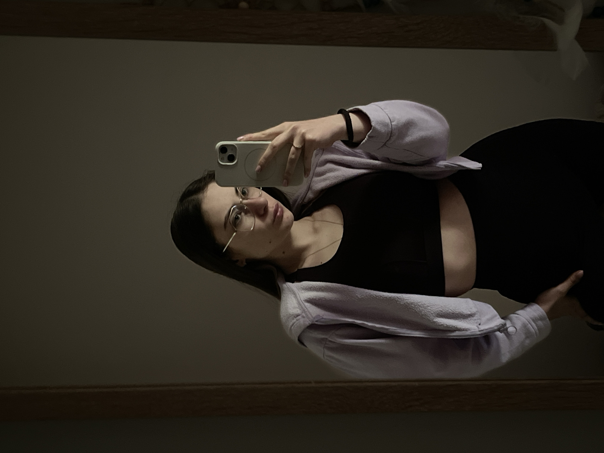 A woman is taking a selfie in front of a mirror. She has long, dark hair and wears glasses. She is wearing a black top and a light purple jacket. She is holding a phone with her right hand and looking at the screen. The background is a dark wall. The location is Unknown, Unknown.