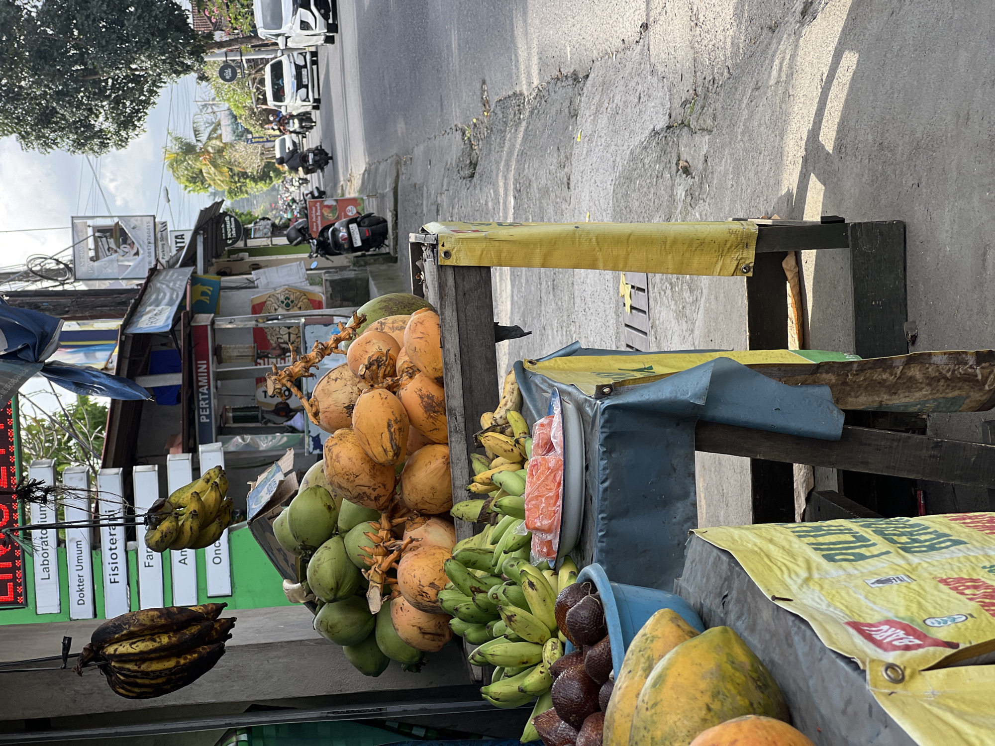 The image captures a vibrant fruit stand on a bustling street in what appears to be the Ubud District of Indonesia. A colorful array of produce is displayed, including bunches of green and yellow bananas, clusters of green and orange coconuts, papayas, and brown salak. The fruit is arranged on a weathered wooden stand, covered with yellow and blue tarps.
In the background, the street teems with activity, including parked motorbikes and passing cars. Signs in Indonesian are visible on nearby buildings, including what appears to be the location of a Laboratory, Physiotherapy, and a Pharmacy. The image is taken at street level, providing an intimate view of the local marketplace.