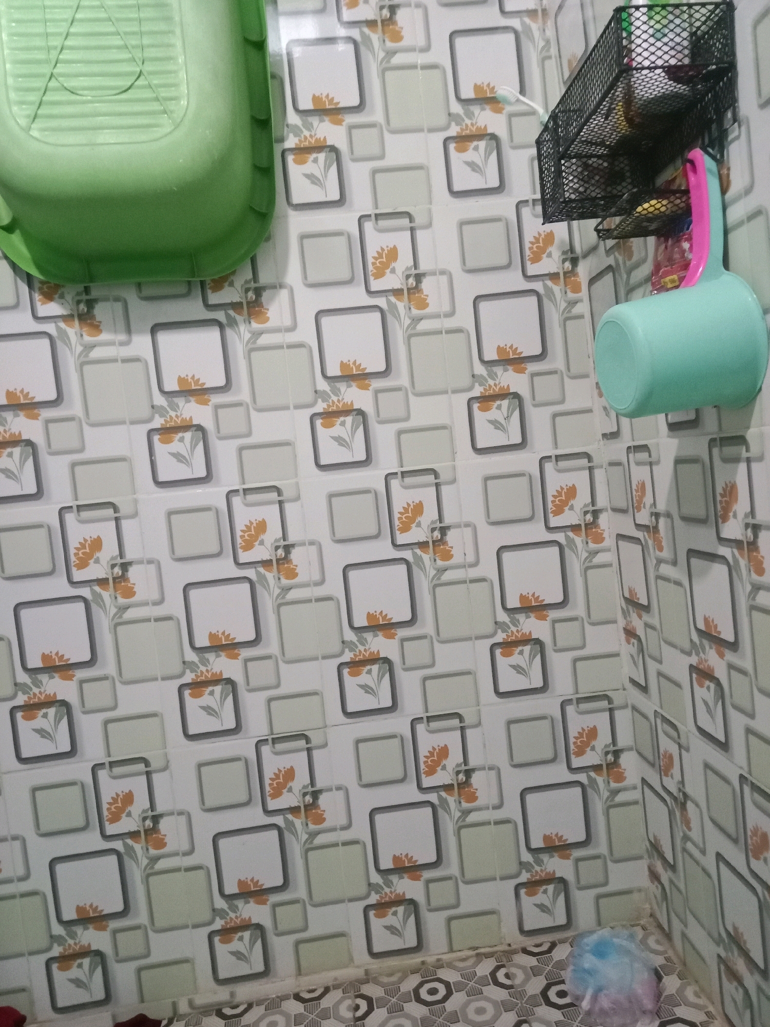 The image depicts a close-up view of a bathroom interior. The walls are covered in a patterned tile featuring squares with grey outlines, some containing floral designs in orange and green, and others left blank or filled with light grey. On the left side of the frame, a bright green plastic basin or tub is visible, its ribbed interior facing upwards.
In the upper right quadrant, a black wire mesh organizer is mounted on the wall, holding various items including what appears to be a toothbrush and possibly toiletries. Hanging from this organizer is a light blue plastic dipper with a pink handle. A white toothbrush is also visible sticking out from the organizer.
The floor is tiled with a pattern of interlocking grey and white geometric shapes. In the bottom right corner of the image, a multi-colored fluffy object, possibly a shower pouf or sponge, rests on the floor. The overall impression is of a functional, if somewhat cluttered, bathroom space. The scene is consistent with a residential bathroom in Gambarsari, Indonesia, given the common tiling and bathroom accessories found in the region. No people are present in the image.
