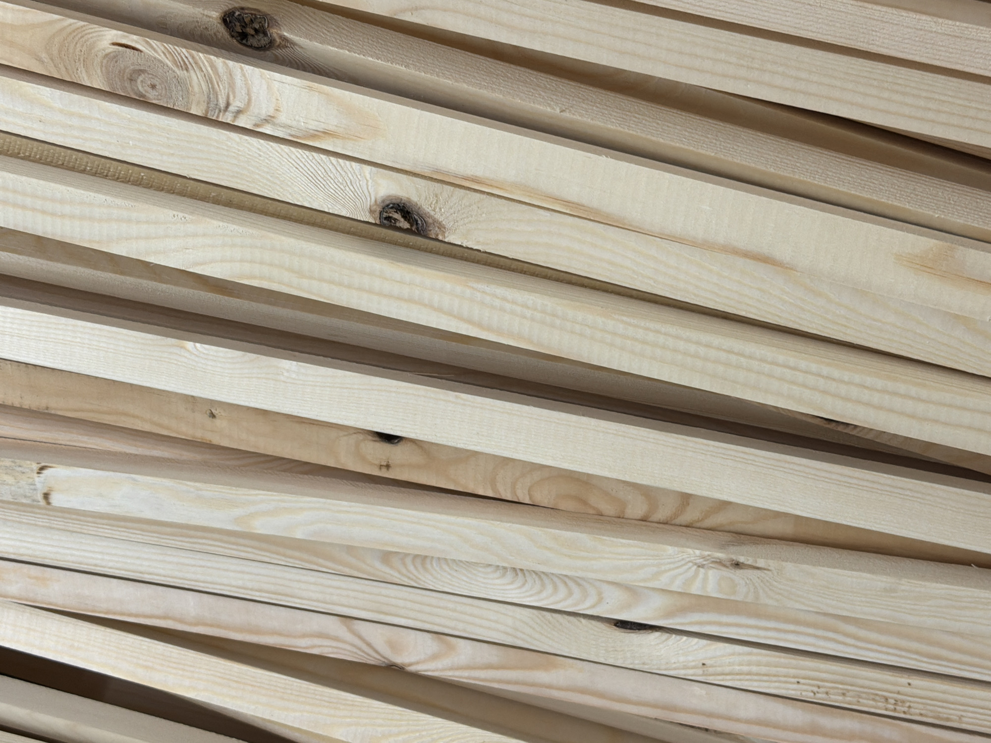 The image shows a stack of light-colored wooden planks. The planks are laid diagonally across the frame, partially overlapping each other. Some planks have knots in the wood. There is no indication of the location being Izhevsk, Russia, or any specific city. The image focuses solely on the stacked wood.