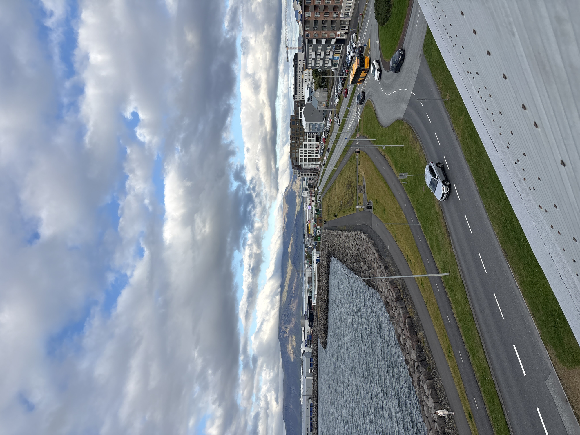 The image depicts a view of the coast and a road in Reykjavik, Iceland, likely taken from an elevated position such as a building or a bridge. In the foreground, a textured, metallic roof surface occupies the bottom right corner. A paved road with white lane markings curves through the scene, with several vehicles including a silver SUV, a white car, a dark sedan, and a yellow bus on it. Adjacent to the road are grassy areas and pathways.
Along the coastline, a breakwater made of large stones lines the edge of a dark, choppy body of water. A paved pathway runs alongside the breakwater, and a single person in a light-colored coat is visible walking on this path. Further inland, a row of buildings, likely commercial or residential, is visible along the roadside, with some industrial structures and cranes in the distance. In the background, a mountain range with a cloudy sky above it forms the horizon. The sky is characterized by a dramatic display of clouds, with patches of blue sky interspersed between them. The overall impression is that of an urban coastal landscape under a cloudy, possibly cool, day.