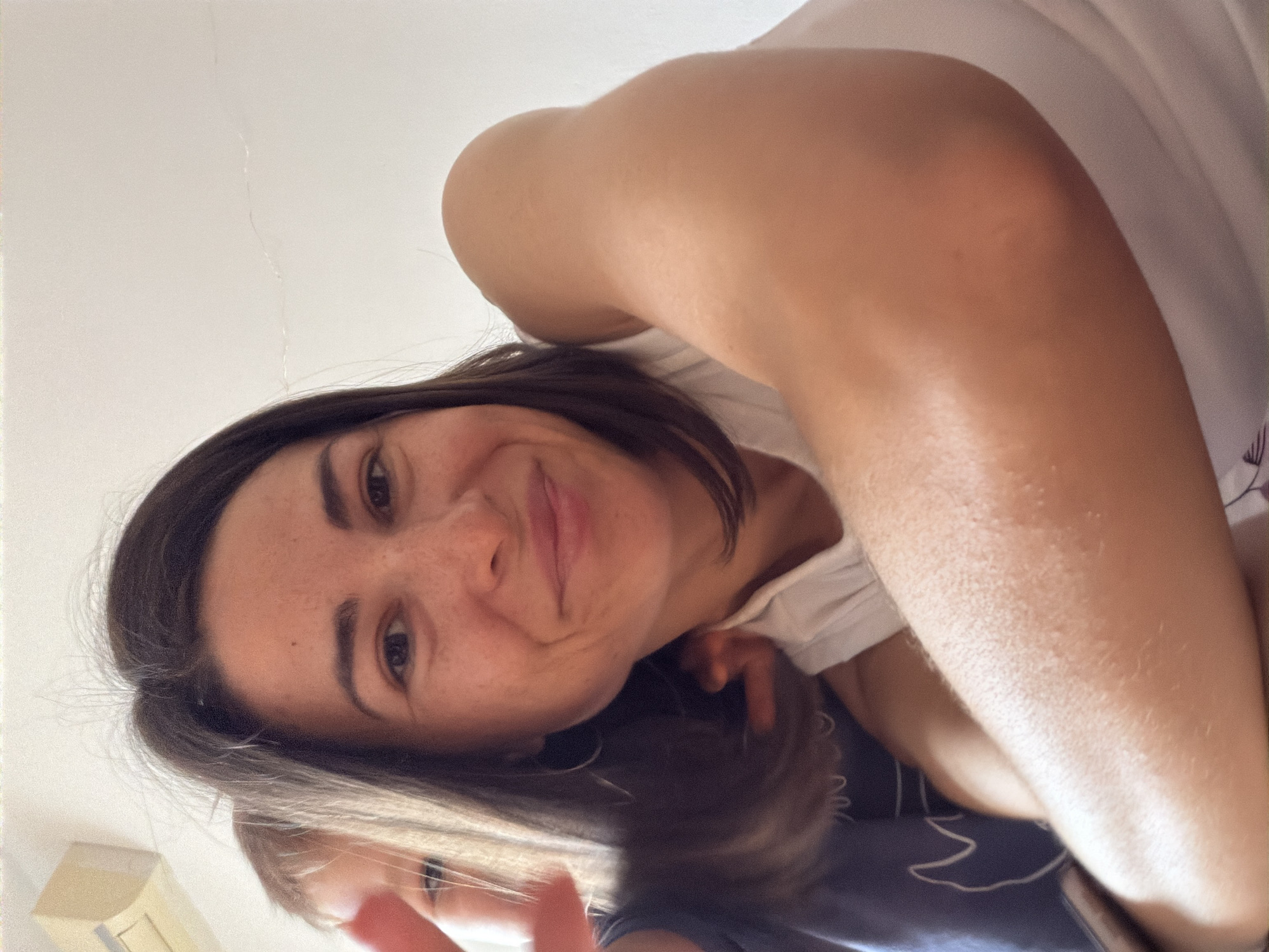 A selfie-style, high-angle shot depicts a woman with shoulder-length dark hair and light skin with freckles. She is smiling at the camera. Parts of her white top are visible along with her arm. Beneath her is a child with blonde hair and fair skin wearing a dark blue shirt. The background is blurred and out of focus, but a white wall is visible in the upper-left corner. The image does not provide enough detail to determine a specific location within the city of Budva, Montenegro.