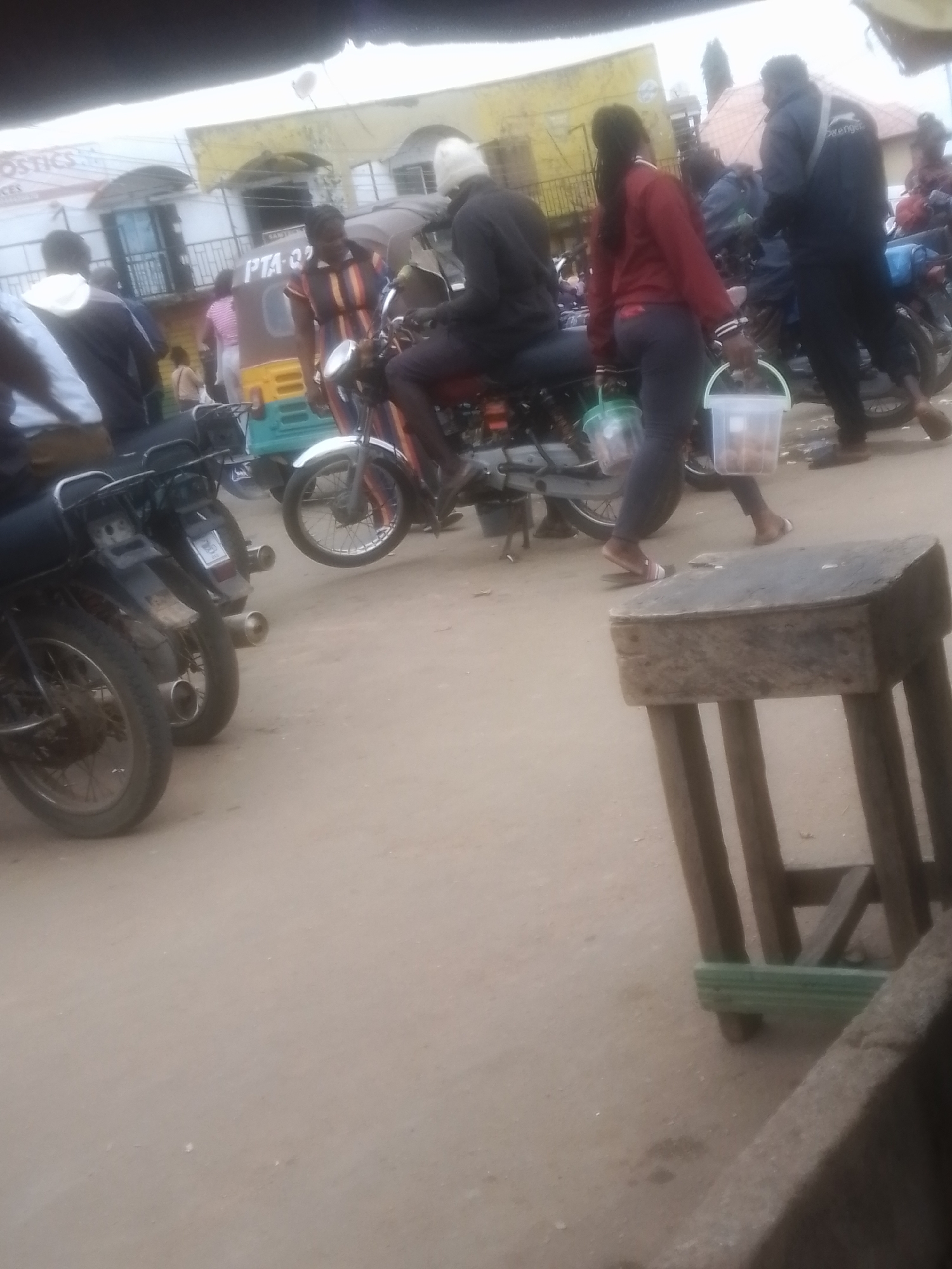 An outdoor, ground-level shot captures a busy street scene. In the foreground, a rudimentary wooden stool with a worn, dark surface sits on a dirt road. Behind it, several motorcycles are parked. One motorcycle on the left has its rear wheel and exhaust pipe clearly visible. Another motorcycle in the center has a rider, a person dressed in dark clothing, with a white cloth wrapped around their head. The rider is seated on the motorcycle, which features a colorful, patterned seat. In the background, a light-colored three-wheeled vehicle, possibly a tuk-tuk, is visible. A person in a striped dress stands near the tuk-tuk. Further in the background, buildings with awnings and signage are present, suggesting a commercial area. Two individuals are walking in the mid-ground, each carrying a plastic bucket. One person, wearing dark pants and a red jacket, holds a white bucket with light-colored contents. The other person, in dark clothing, carries a green bucket. The overall impression is one of a bustling market or street corner.