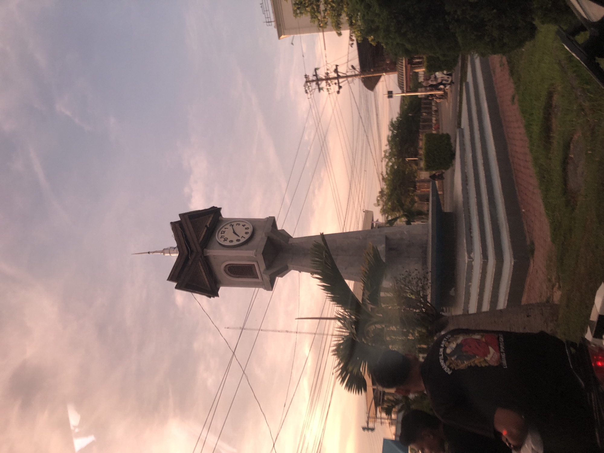 A photo of the clock tower in Lhokseumawe, Indonesia, during sunset. The clock is in the center of the frame, with the sky behind it and a palm tree slightly obstructing it. People are visible in the foreground. Power lines are going from the upper left to the right. Steps can be seen leading away from the clock tower. Grass and trees are on the right.