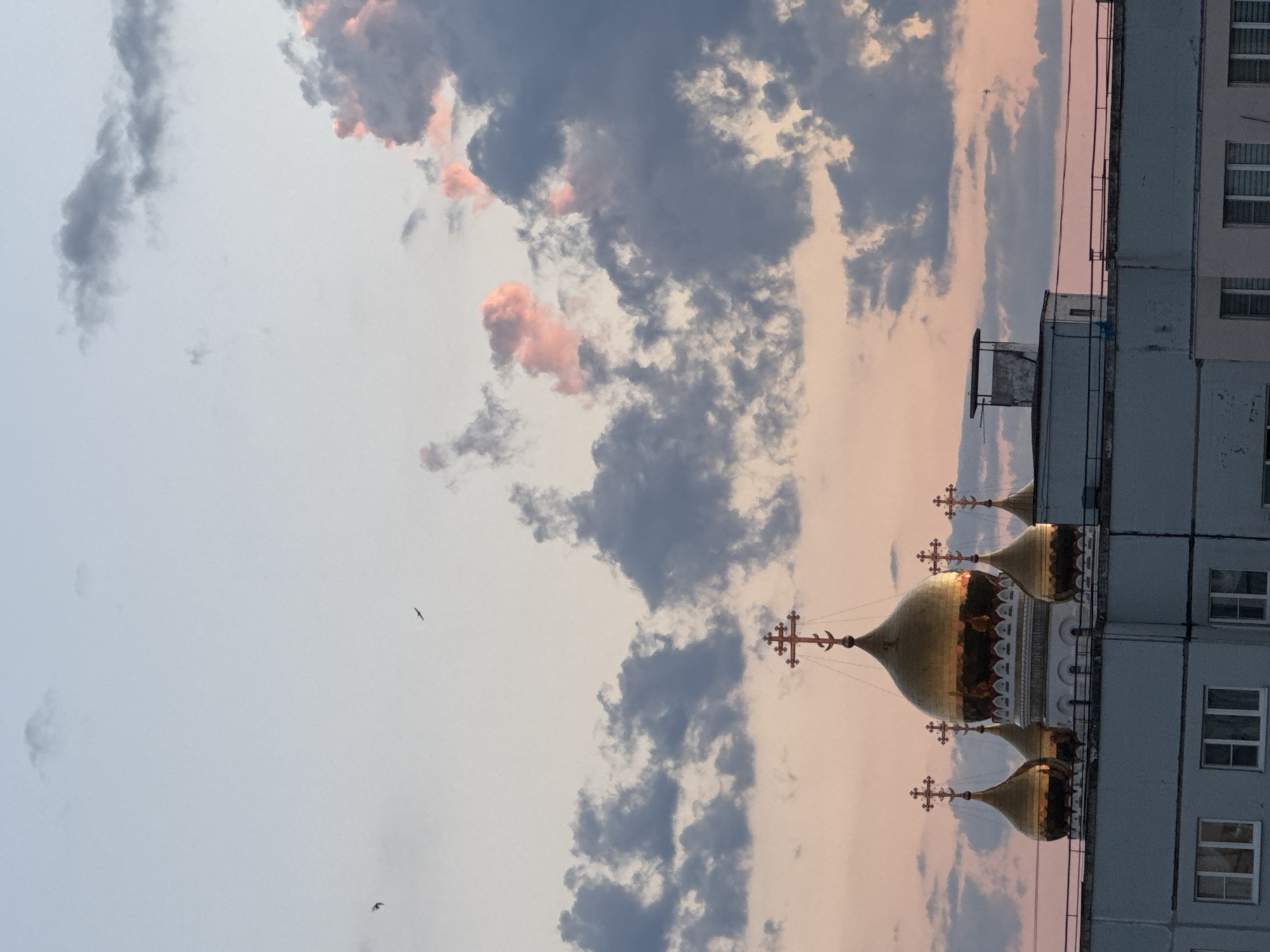 The image shows an Orthodox church with golden domes and crosses, seen from a low angle behind a building. The sky is filled with clouds, with some illuminated by pink and orange hues, suggesting a sunset or sunrise. Two birds are visible in the sky, appearing as small silhouettes against the lighter background. The building in the foreground is a multi-story structure with windows, suggesting an urban environment. The scene is peaceful and serene, with the soft light and the prominent religious architecture creating a sense of spiritual contemplation. The overall impression is one of beauty and tranquility.