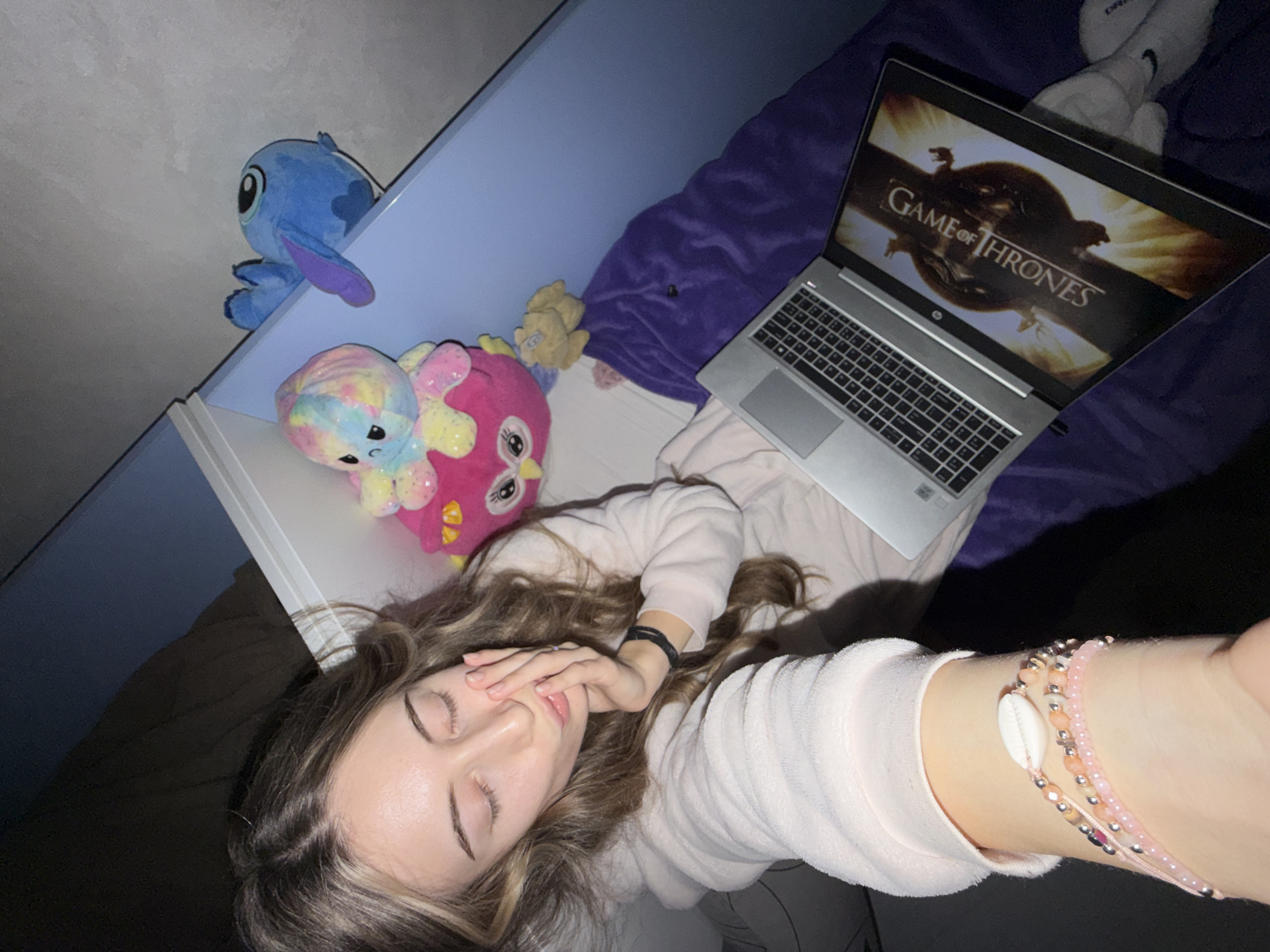 A woman lies on a bed with a laptop showing "Game of Thrones." She wears a light pink sweatshirt and bracelets. To her left are stuffed animals, including Stitch. The environment appears to be a bedroom. Location is unknown, and there is no indication it is Plovdiv, Bulgaria.