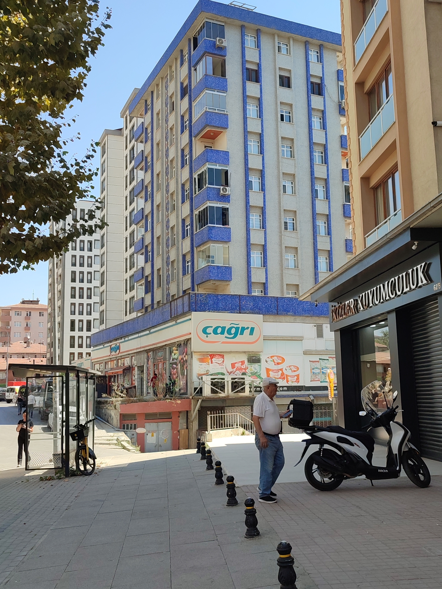 The image captures a street scene in Kadıköy, Turkey, likely on a sunny day, with a clear blue sky. The focal point is a multi-story apartment building with a striking facade featuring blue accents and numerous windows. To the right of this building is another structure, an apartment building, with a beige exterior and balconies with glass railings.
At the ground level of the blue-accented building, there are storefronts. One prominent sign reads "çağrı" in white text on a blue and orange background, suggesting a business such as a market or grocery store. Adjacent to it, to the right, is a storefront with a dark facade and a sign that says "KIZILAR KUYUMCULUK," indicating a jewelry store.
In the foreground, a man wearing a white polo shirt and blue jeans stands on the sidewalk, holding a black bag. To his right, a white and black scooter with a transparent windshield is parked. The sidewalk is paved with gray tiles and bordered by small black bollards topped with yellow accents. A bus stop shelter with glass panels is visible on the left side of the image, with a bicycle parked inside. A woman in black clothing stands near the bus stop. In the background, other buildings and trees can be seen, contributing to the urban environment. The image is a street-level perspective, looking up at the buildings and capturing daily urban life.