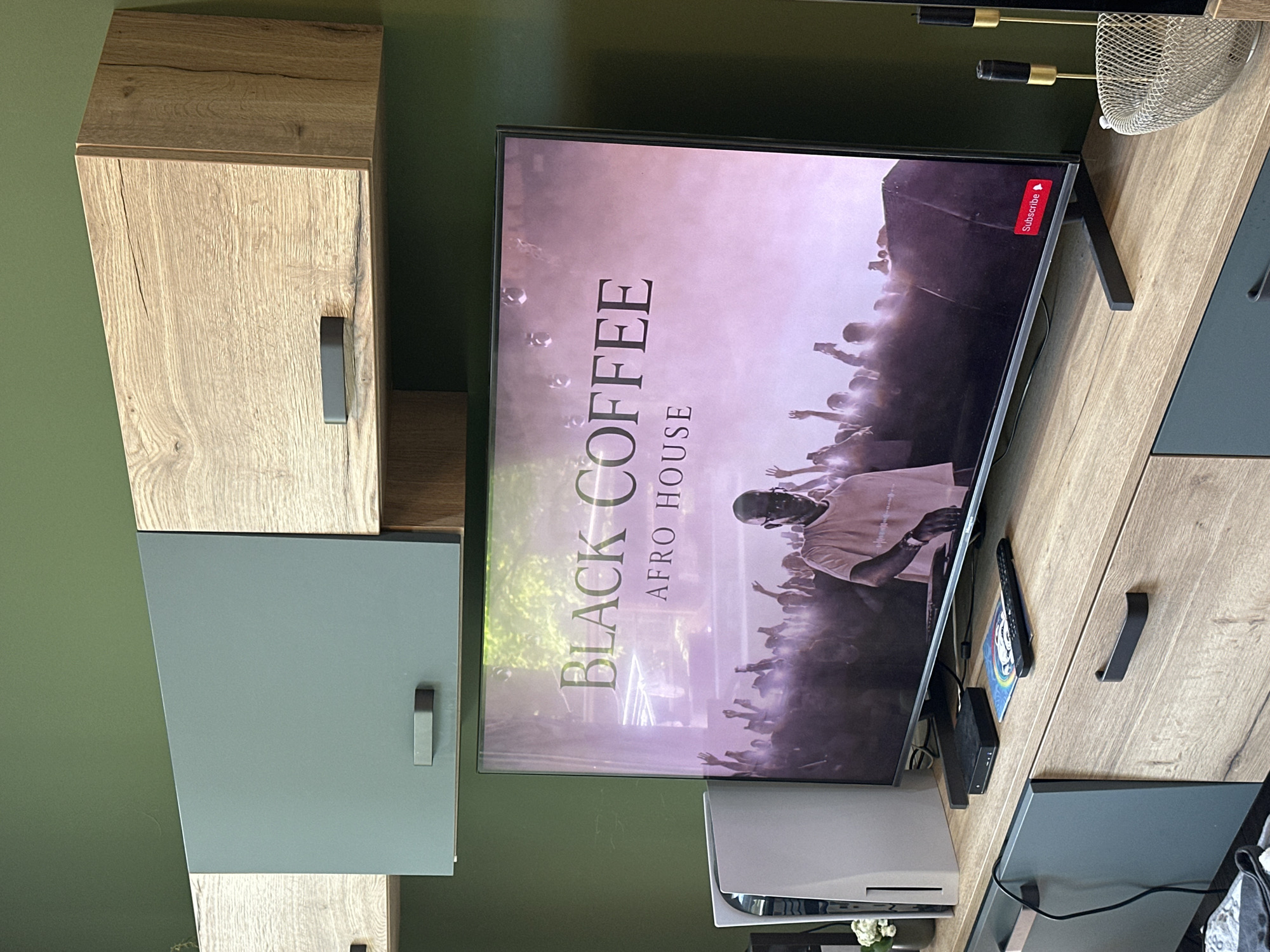 An indoor scene captures a modern entertainment setup, likely in a living room, with a distinct olive green wall serving as the backdrop. The central focus is a large, flat-screen television displaying content. The screen shows a musical event or video featuring "BLACK COFFEE" and the genre "AFRO HOUSE" in prominent white text. Below the text, a DJ (possibly Black Coffee himself) is visible, performing in front of a cheering crowd, illuminated by what appears to be stage lighting or haze. A red "Subscribe" button with a bell icon is visible in the top right corner of the TV screen, suggesting a streaming platform like YouTube.
The TV is part of a contemporary media console that features a combination of light wood-grain panels and sleek, matte grey/teal sections. Dark grey rectangular handles are visible on some of the cabinet doors or drawers. Below the TV, a white PlayStation 5 console stands upright. A black remote control and a small black electronic device (possibly a streaming box or set-top box) are also on the wooden surface next to the TV. Above the TV, a decorative metallic mesh basket and two metallic (gold and black) accent pieces are affixed to the wall or a shelf. The overall aesthetic is modern and clean. The scene is set in Sofia, Bulgaria.